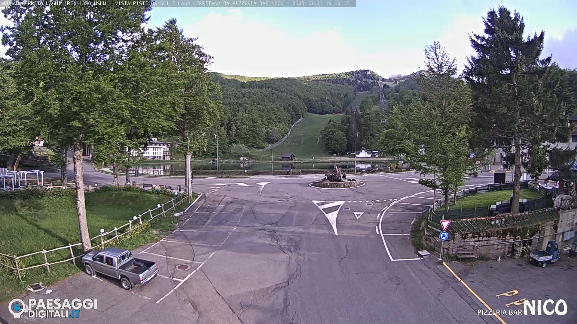 A mostly empty parking lot and roadway intersection are situated next to a small pond, with a forested hillside and ski lift visible in the background under a partly sunny sky.
