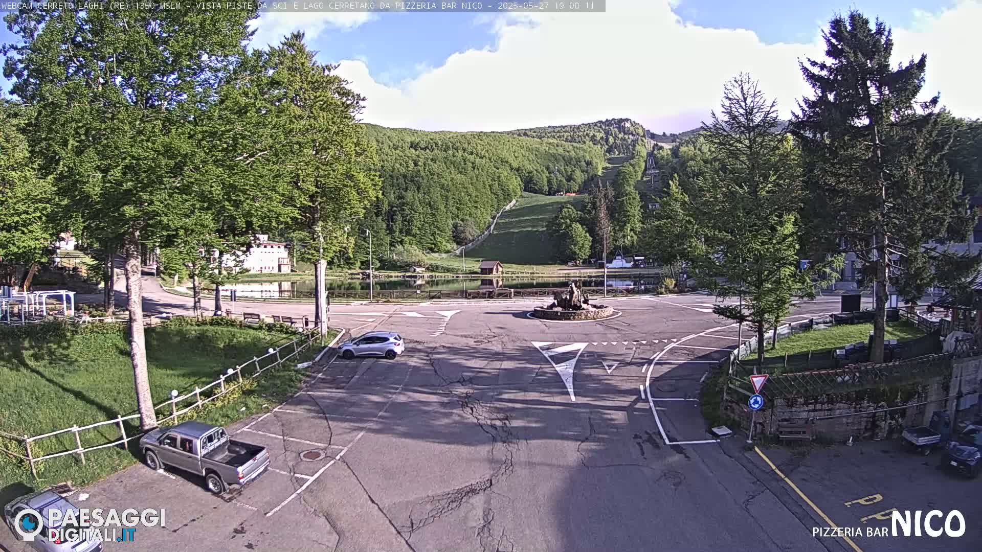 A mostly sunny view of a parking lot and roundabout near a small lake and forested hillside.