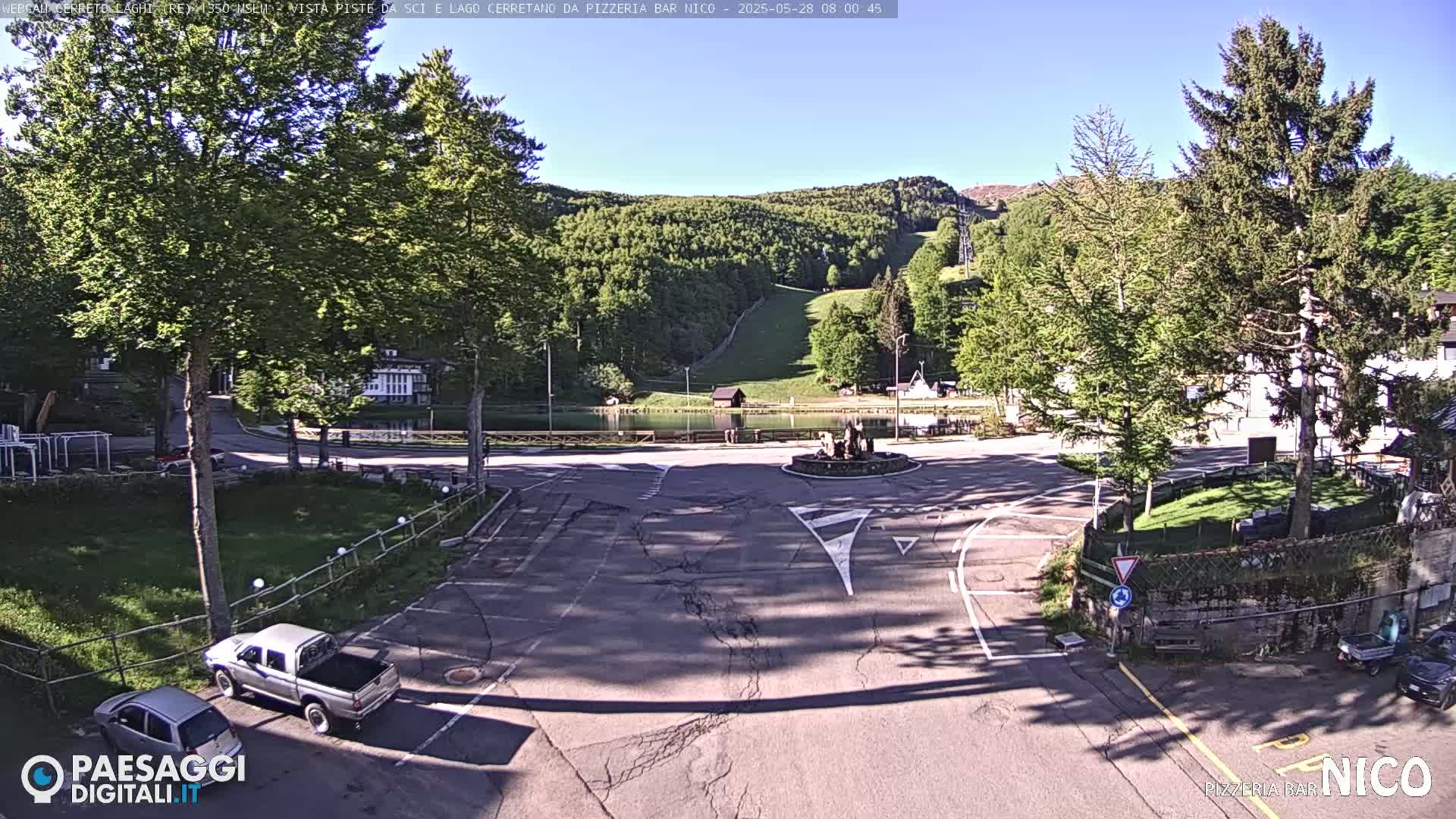 A sunny day shows a parking lot and roundabout near a calm lake, nestled among green, forested hills with a ski slope visible in the distance.