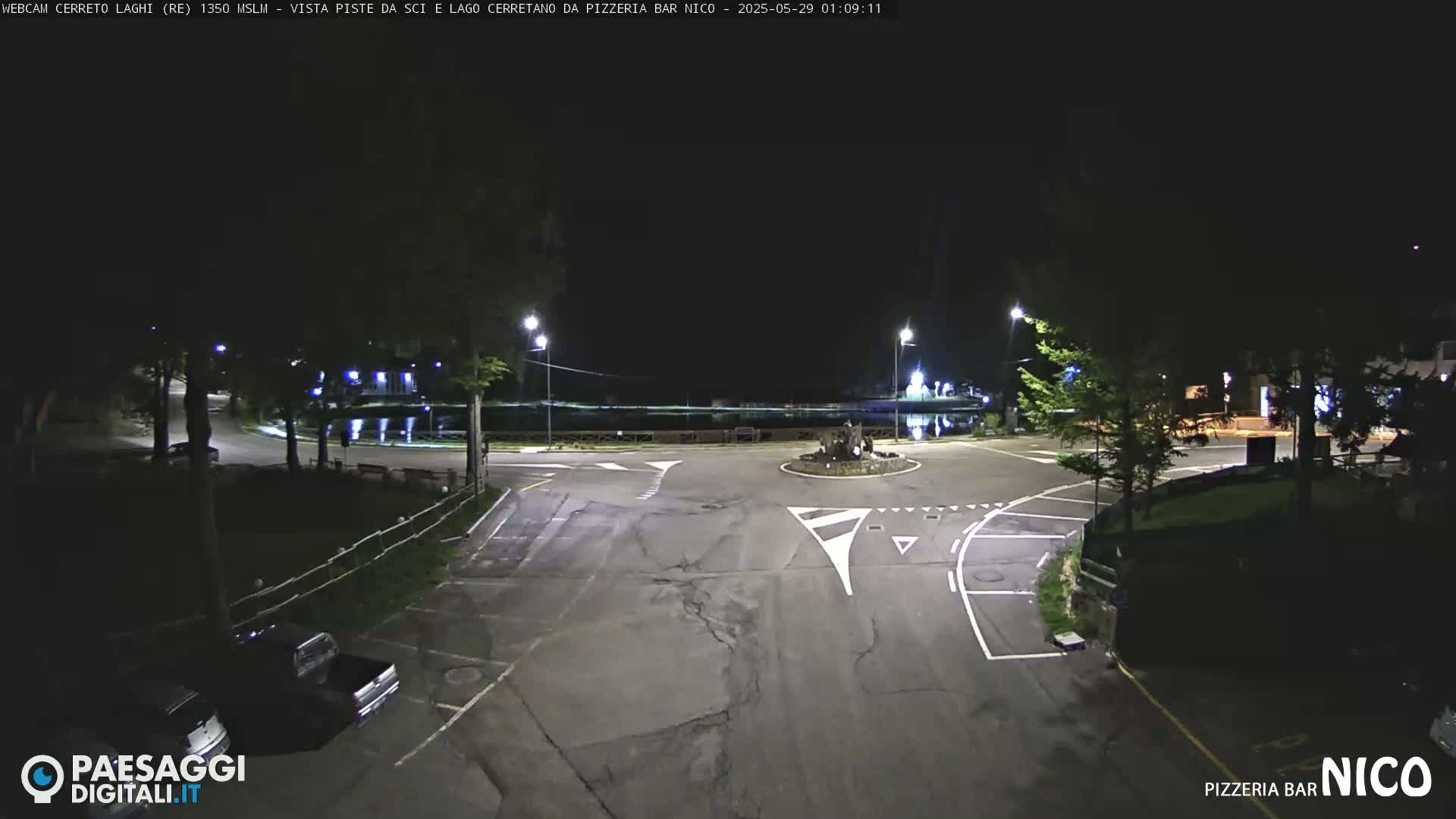 A nighttime view of an empty parking lot and road intersection near a body of water, illuminated by streetlights.