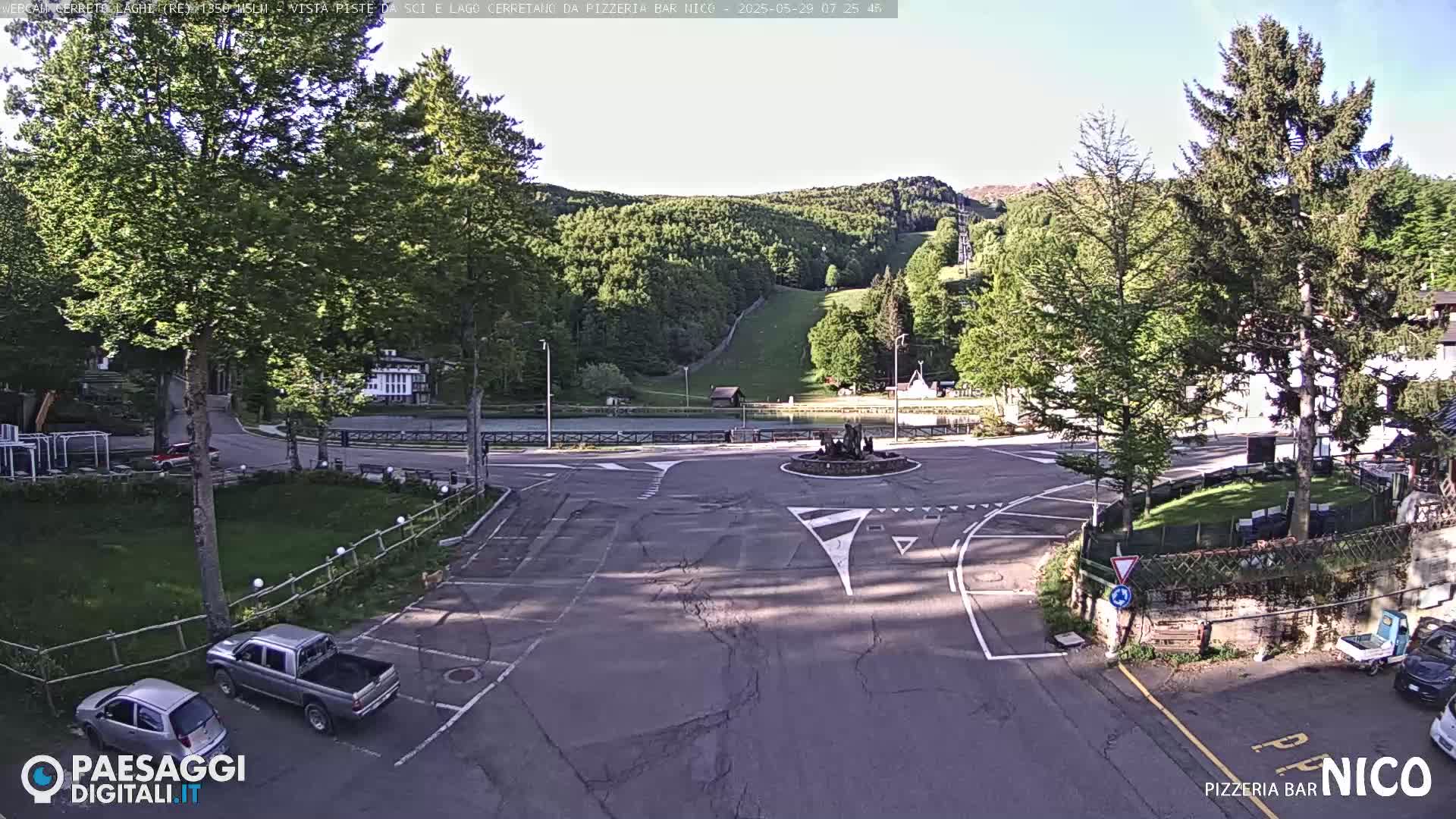 A sunny day reveals a paved intersection in a mountain town, with a few cars parked nearby and a grassy area, with a background of forested hills and a ski slope.