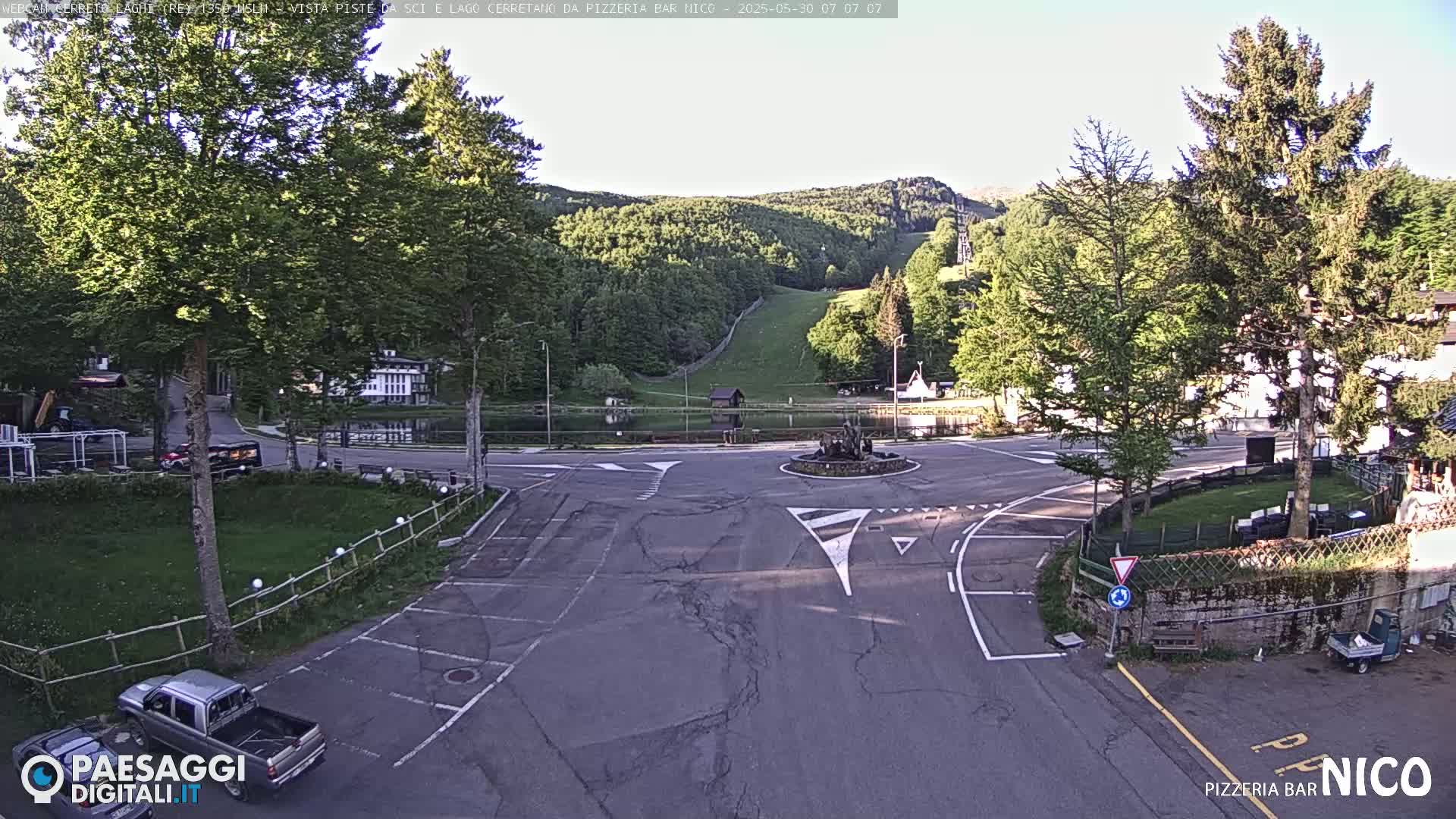 A sunny day reveals a parking area and roundabout near a lake and ski slope, surrounded by lush green trees and buildings.