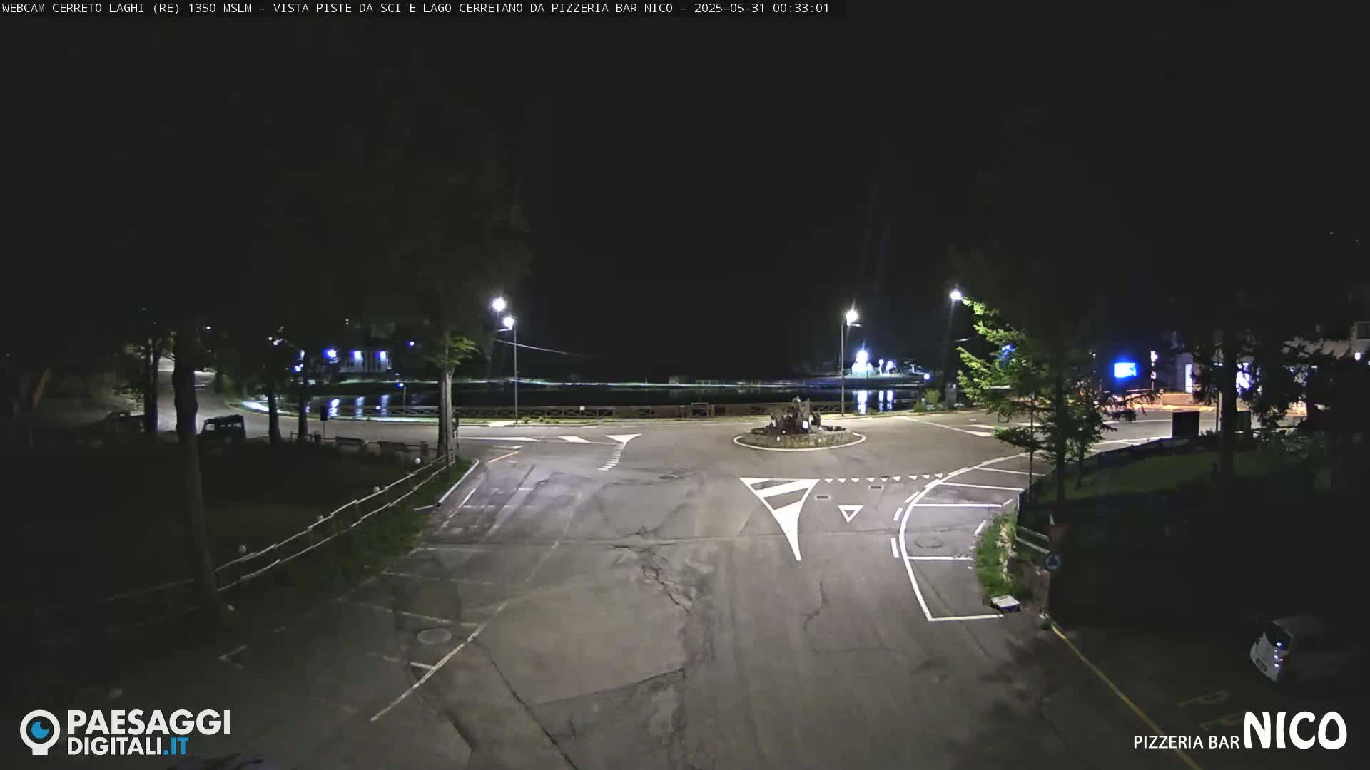 A nighttime view of a dark, empty roundabout near a body of water, illuminated by streetlights.