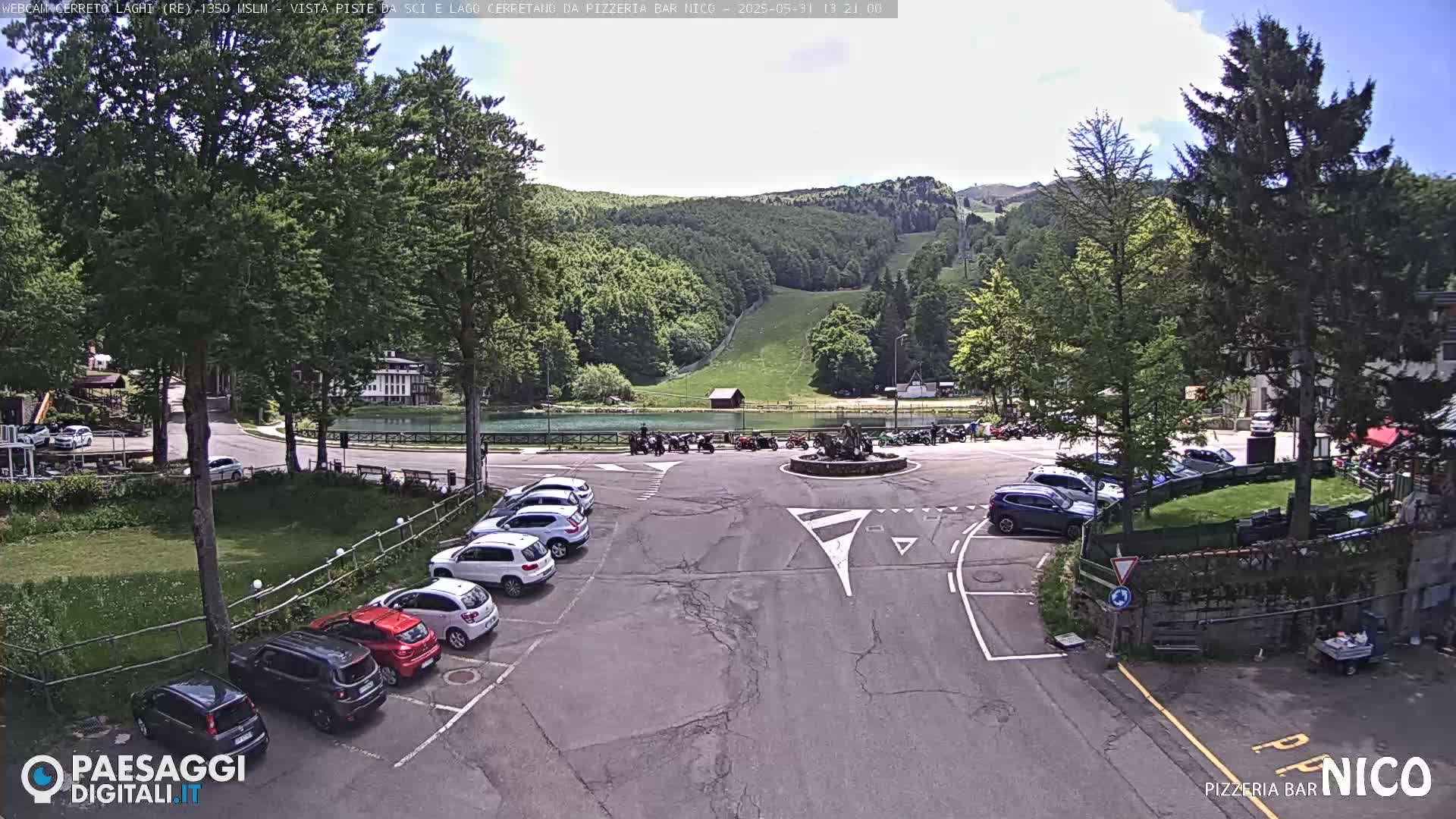 A parking lot with cars is situated in front of a small lake and a ski slope on a sunny day.