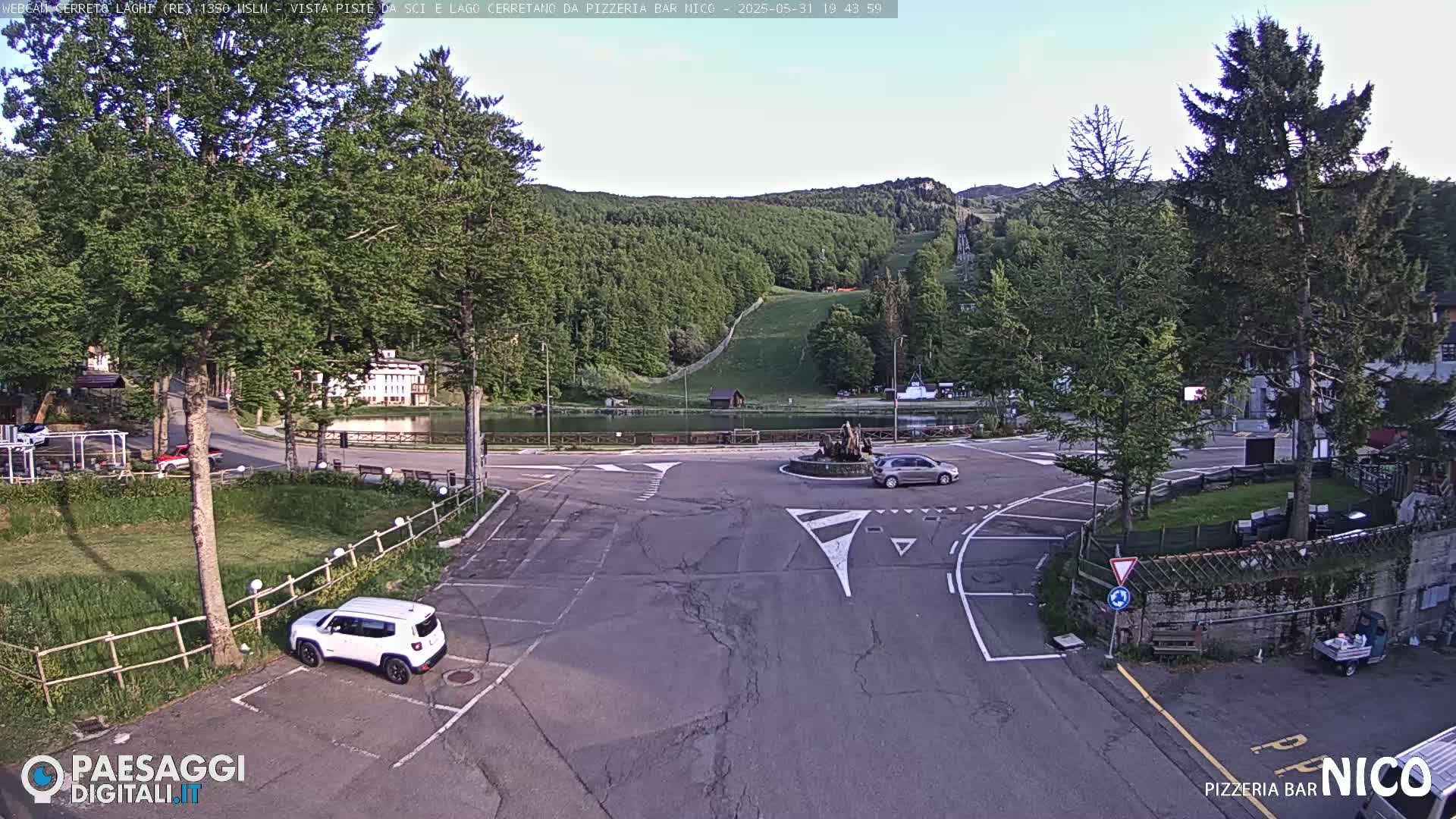 A mostly sunny view of a parking lot, road intersection, and small lake, surrounded by trees and a hillside with a ski slope.