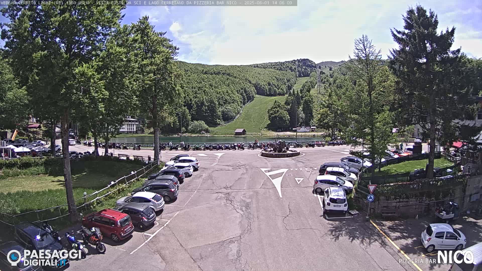 A parking lot filled with cars is situated near a small lake and a grassy hillside with a ski slope under a partly sunny sky.