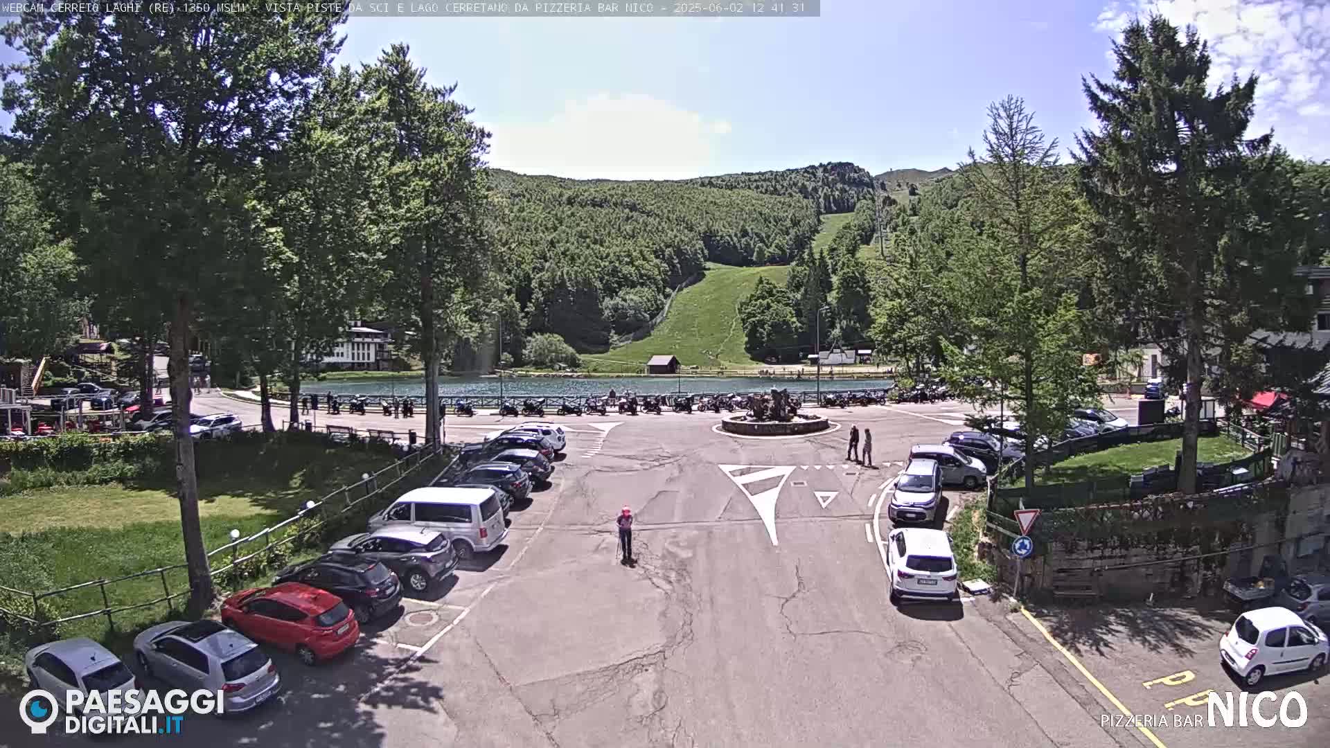 A sunny day shows a parking lot full of cars near a small lake and a hillside with a ski slope, surrounded by lush green trees.