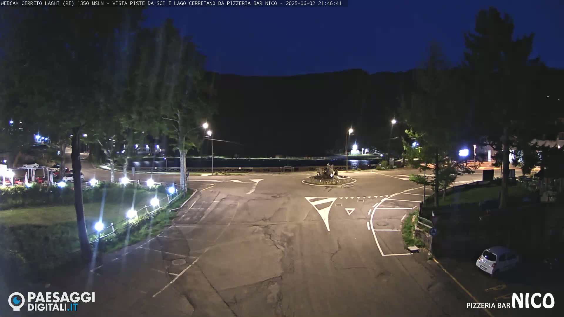 An empty parking lot and roadway at night, illuminated by streetlights, are situated near a calm body of water under a dark, clear sky.