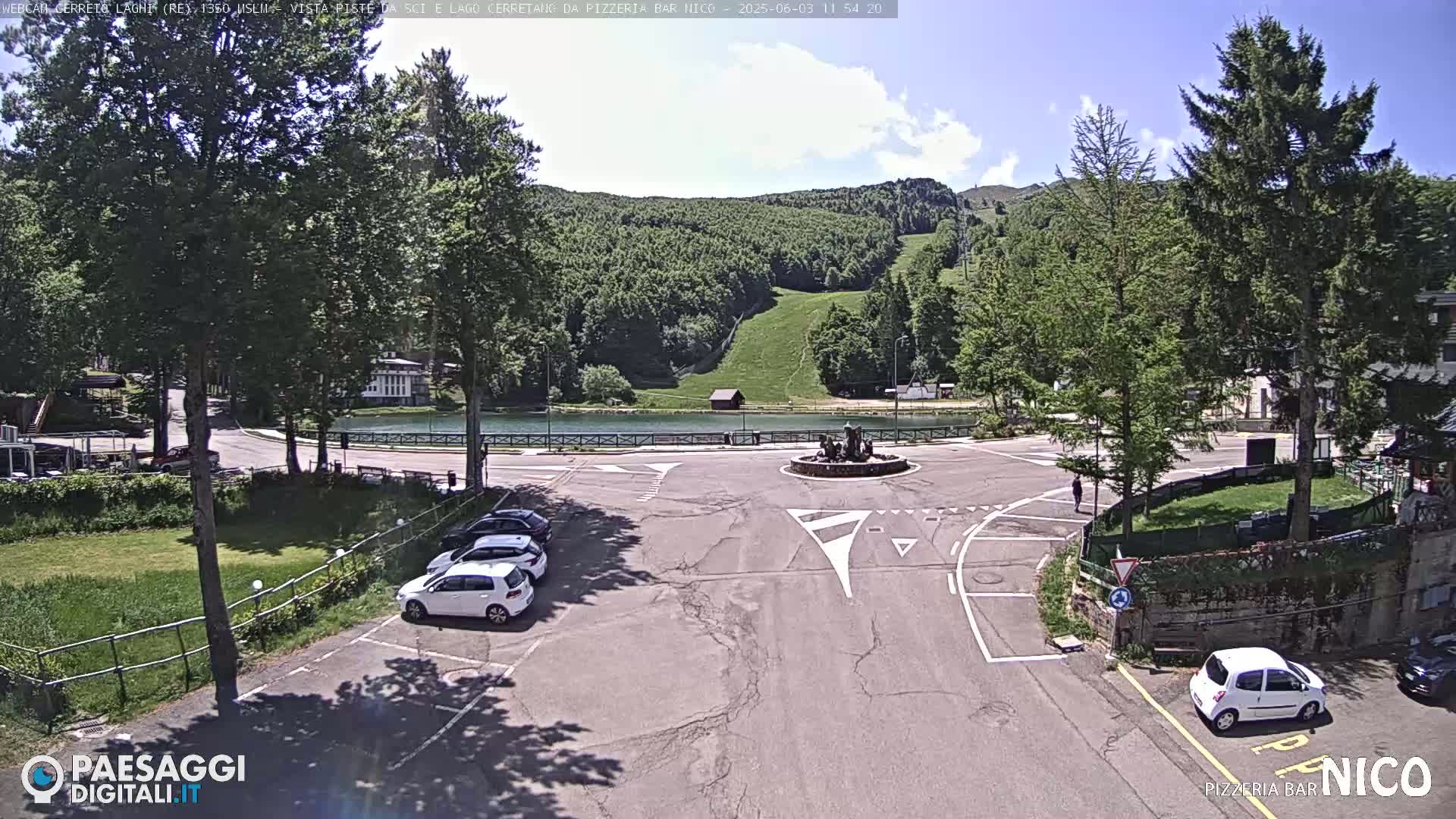 A sunny day reveals a parking lot with several cars, a small pond, and a grassy area surrounded by trees and a hillside with a ski slope in the background.