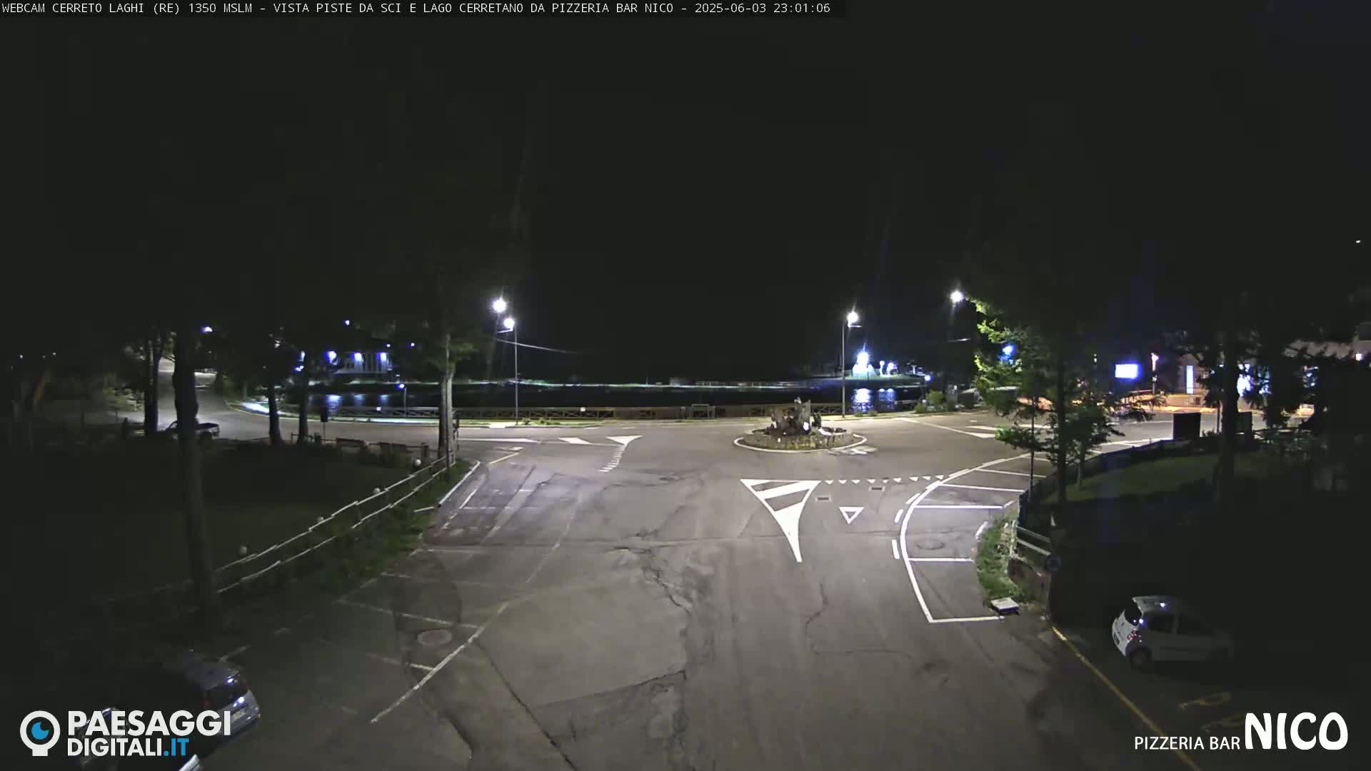 A nighttime view of a mostly empty paved intersection near a body of water, with a few cars parked and streetlights illuminating the scene.