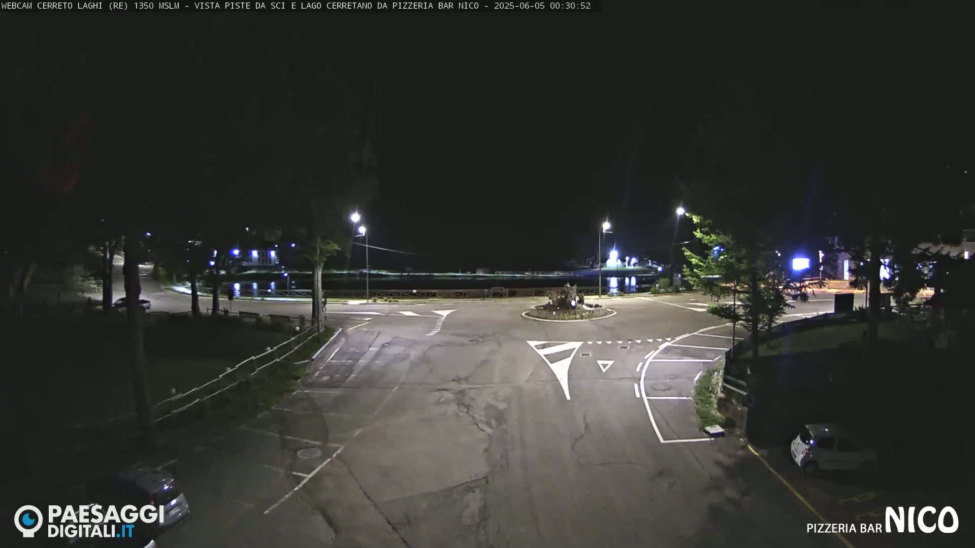 A nighttime view of a mostly empty parking lot and roundabout near a body of water, illuminated by streetlights.