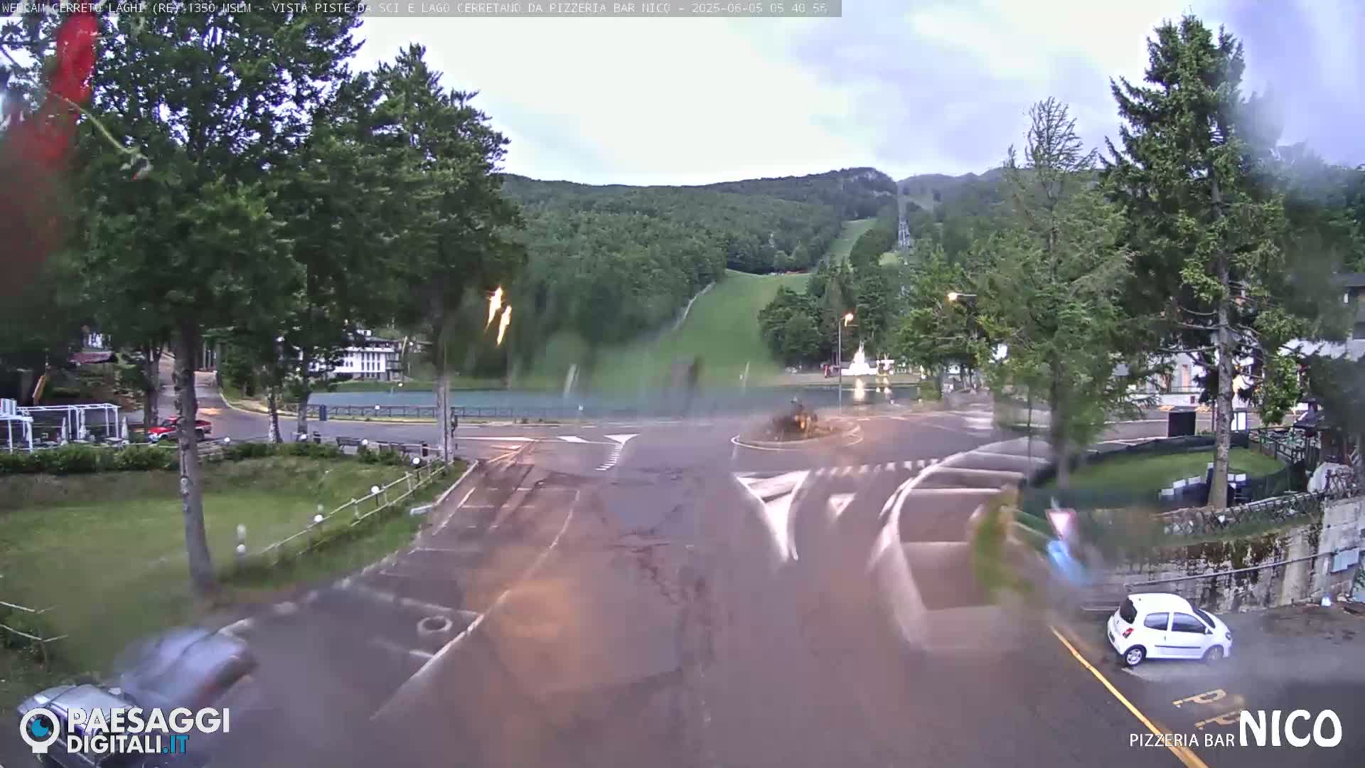 Rain falls on a roundabout near a wooded hillside with ski slopes visible in the distance, as cars drive through the intersection.