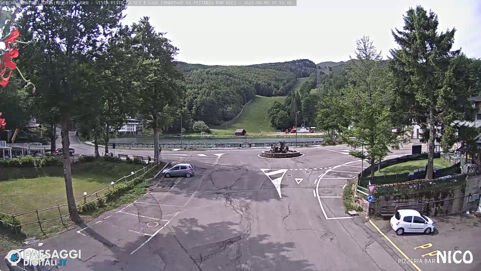 A mostly sunny outdoor scene shows a parking lot and roadway intersection near a lake and ski slope in a mountainous area.