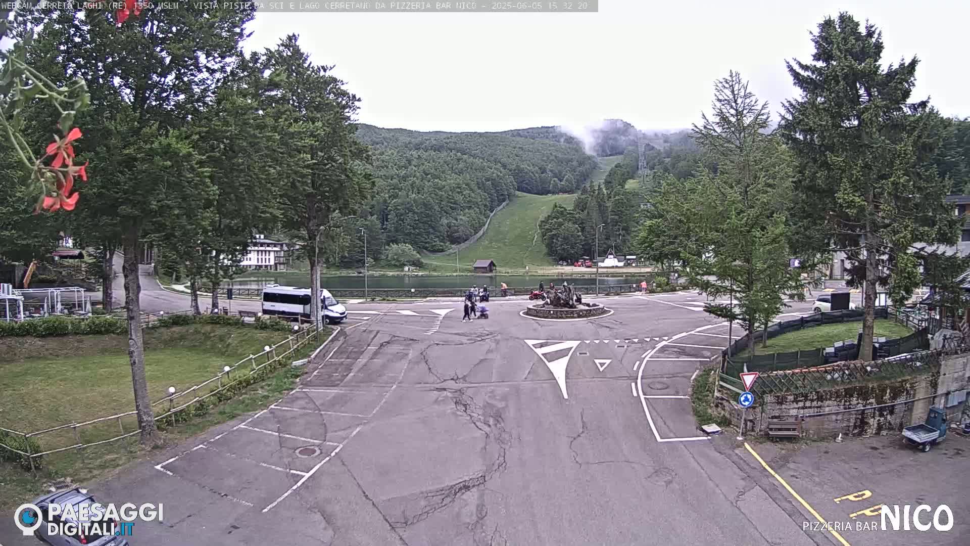 A parking lot and roundabout in a mountainous area with overcast skies, a small lake, and a ski slope visible in the background.