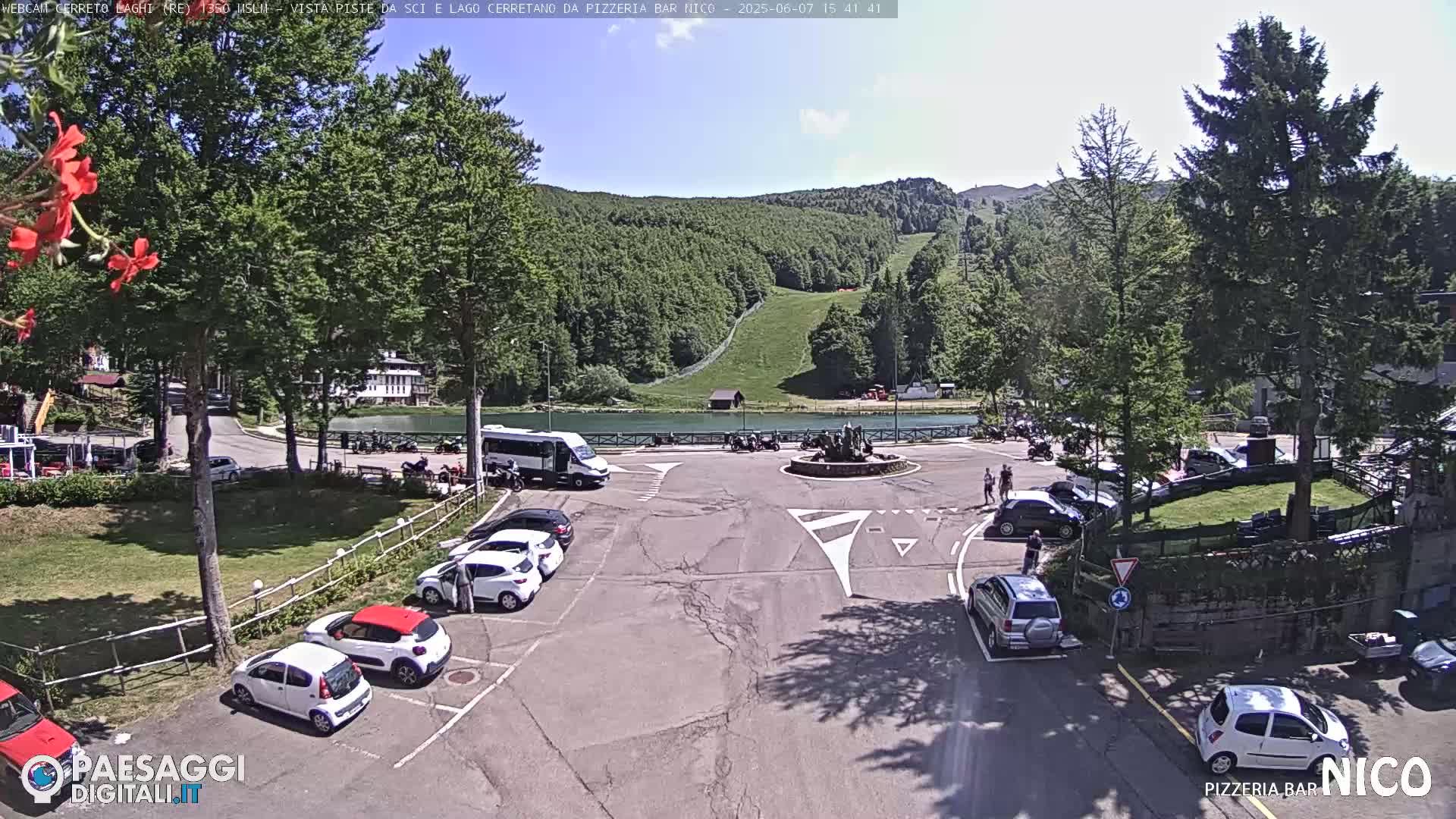 A parking lot with several cars is situated near a small lake and a grassy hill under a sunny sky.