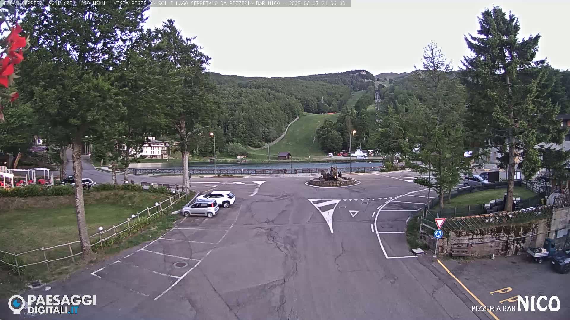 A mostly empty parking lot and roundabout sit in front of a lake and a wooded hillside with a ski slope visible in the distance under an overcast sky.