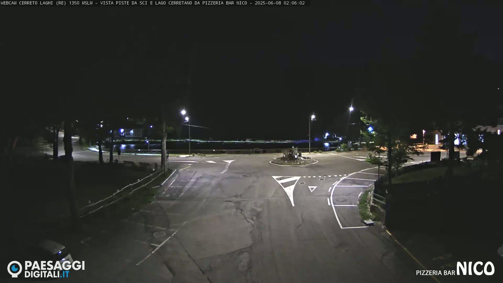 A nighttime view of a dark, empty parking lot and roadway intersection near a body of water, illuminated by streetlights.