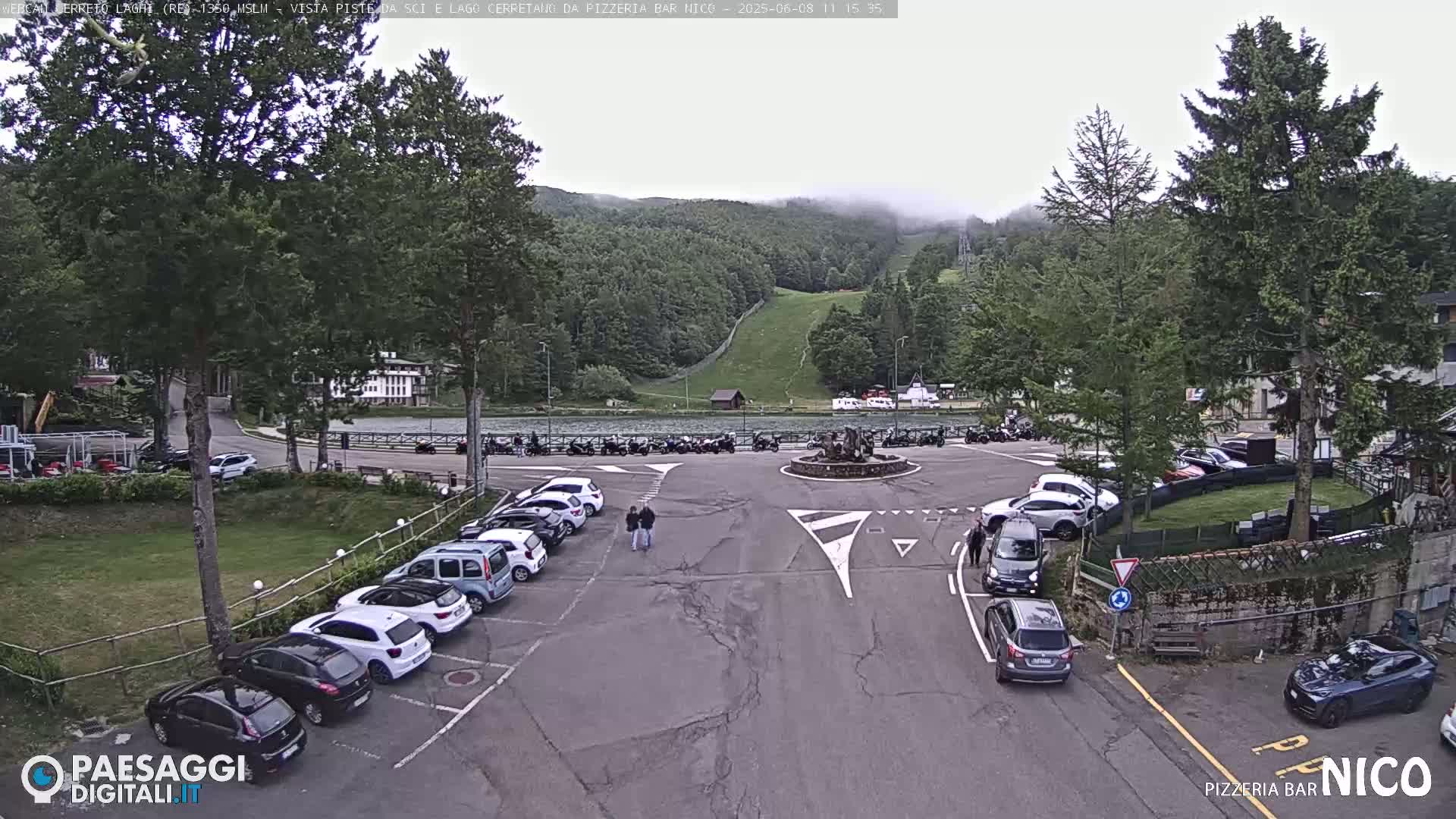 A parking lot with several cars is situated in front of a lake and a wooded hillside under an overcast sky.