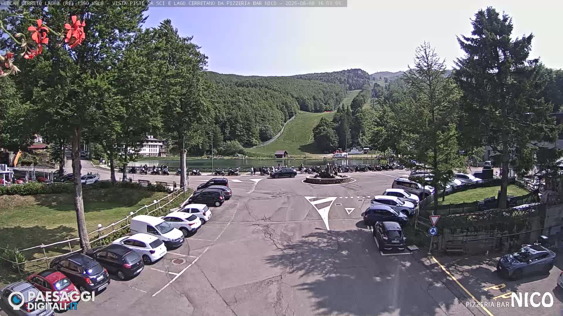 A sunny day at a mountain lake with a parking lot full of cars, a small town in the background, and a grassy hill with a ski lift visible in the distance.