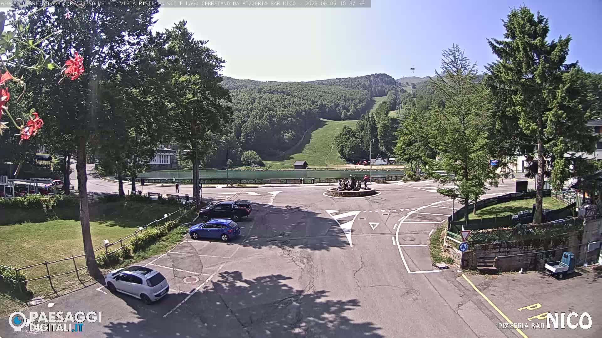 A sunny parking lot with several cars is situated near a small lake and green hills, with a roundabout in the center.
