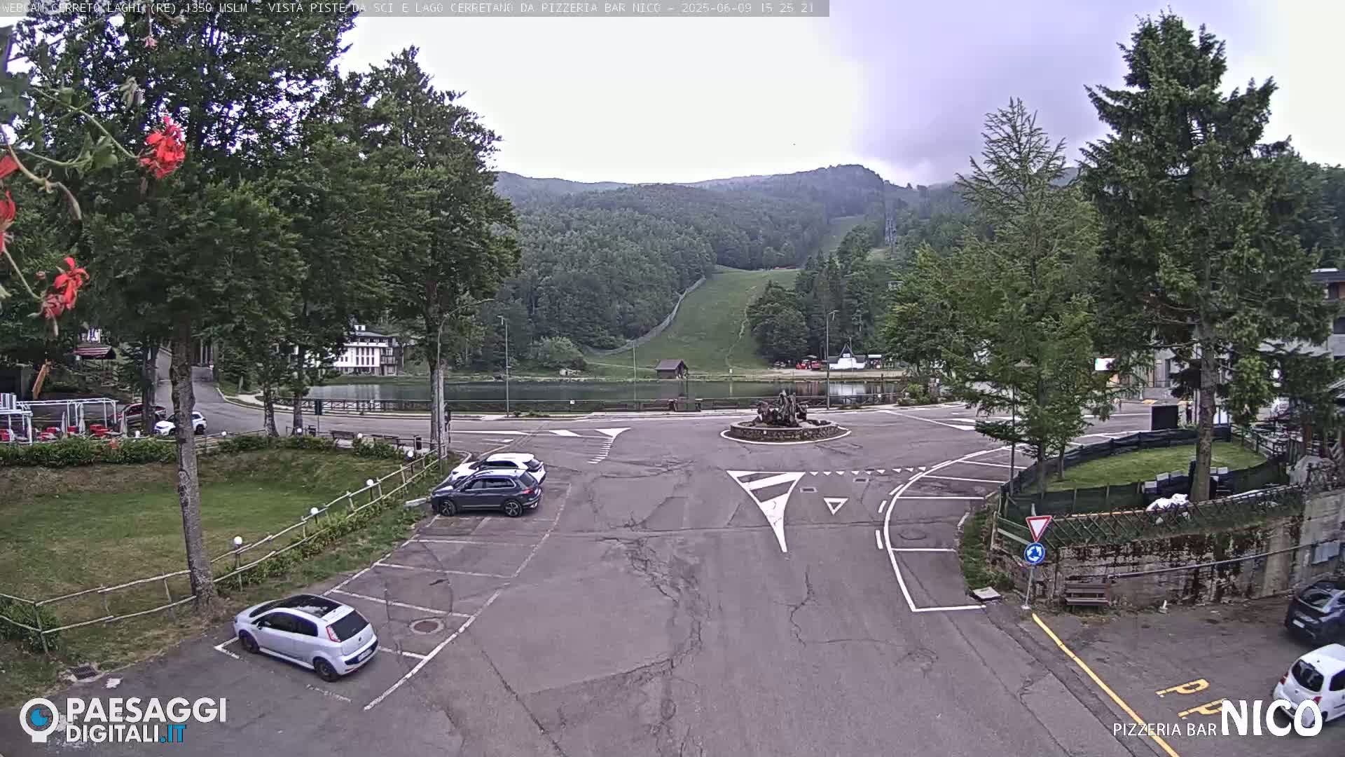 A mostly overcast day shows a parking lot with several cars, a small pond, and a grassy hill with a ski slope in the background.