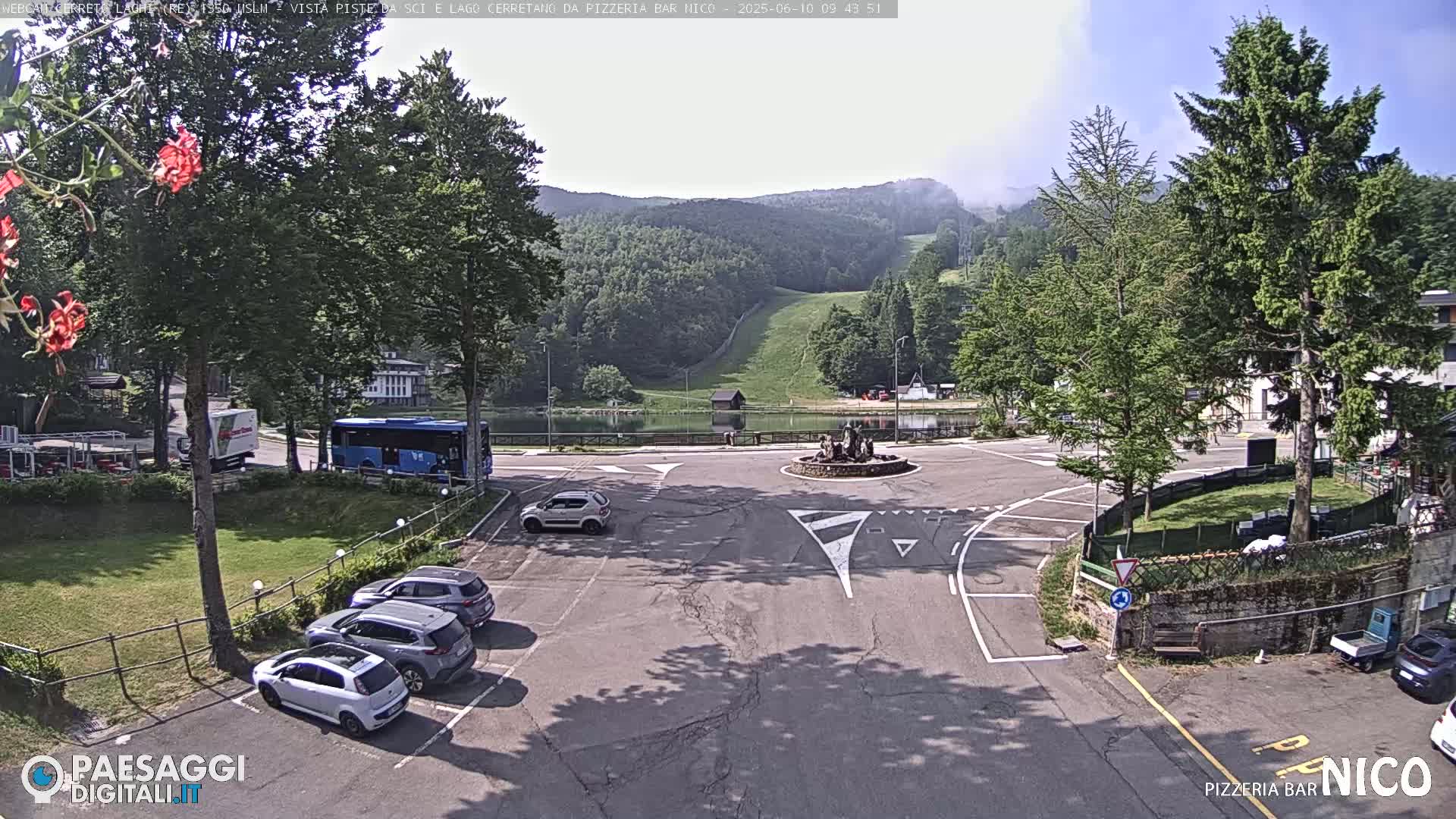 A parking lot with several cars, a bus, and a roundabout in the foreground, overlooks a small lake and a grassy hillside with a ski lift in the background, on a partly sunny day.