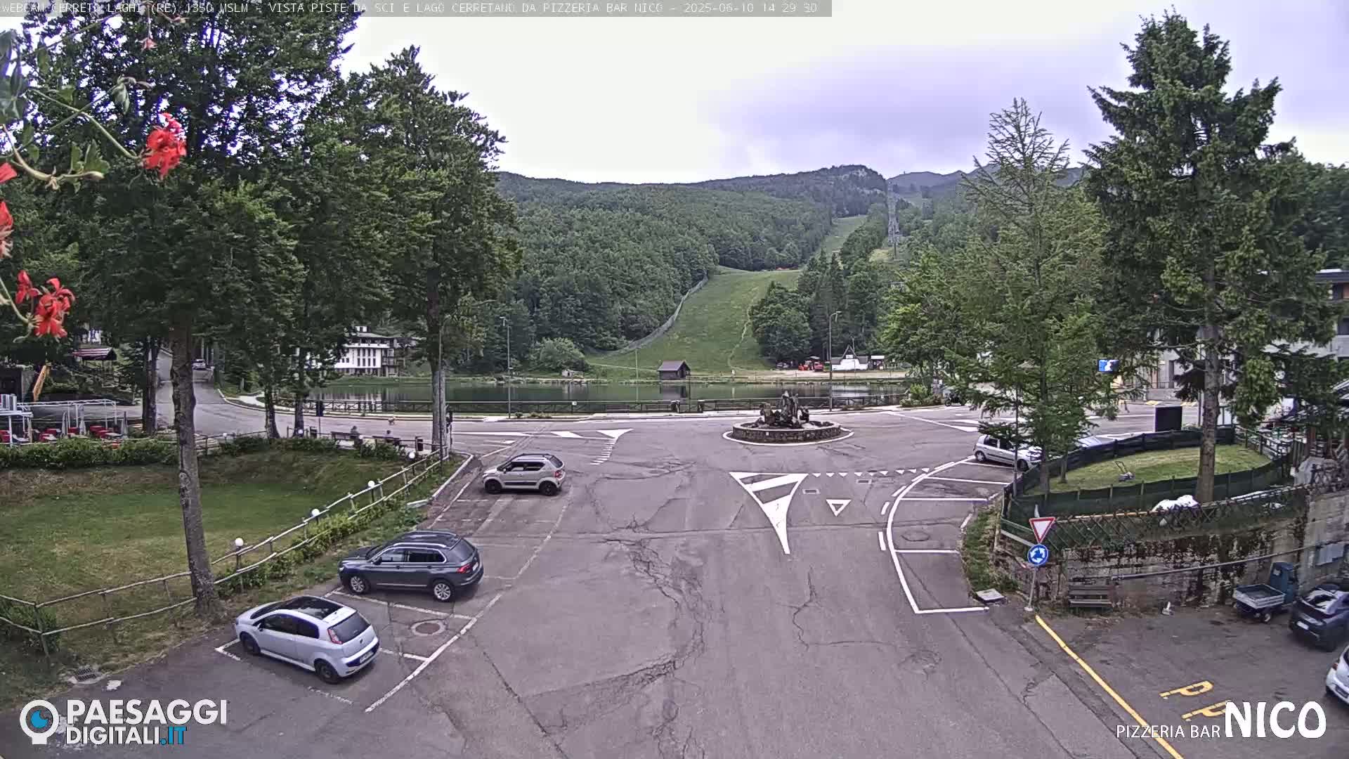 A parking lot with several cars is situated near a calm lake and a ski slope, under an overcast sky.