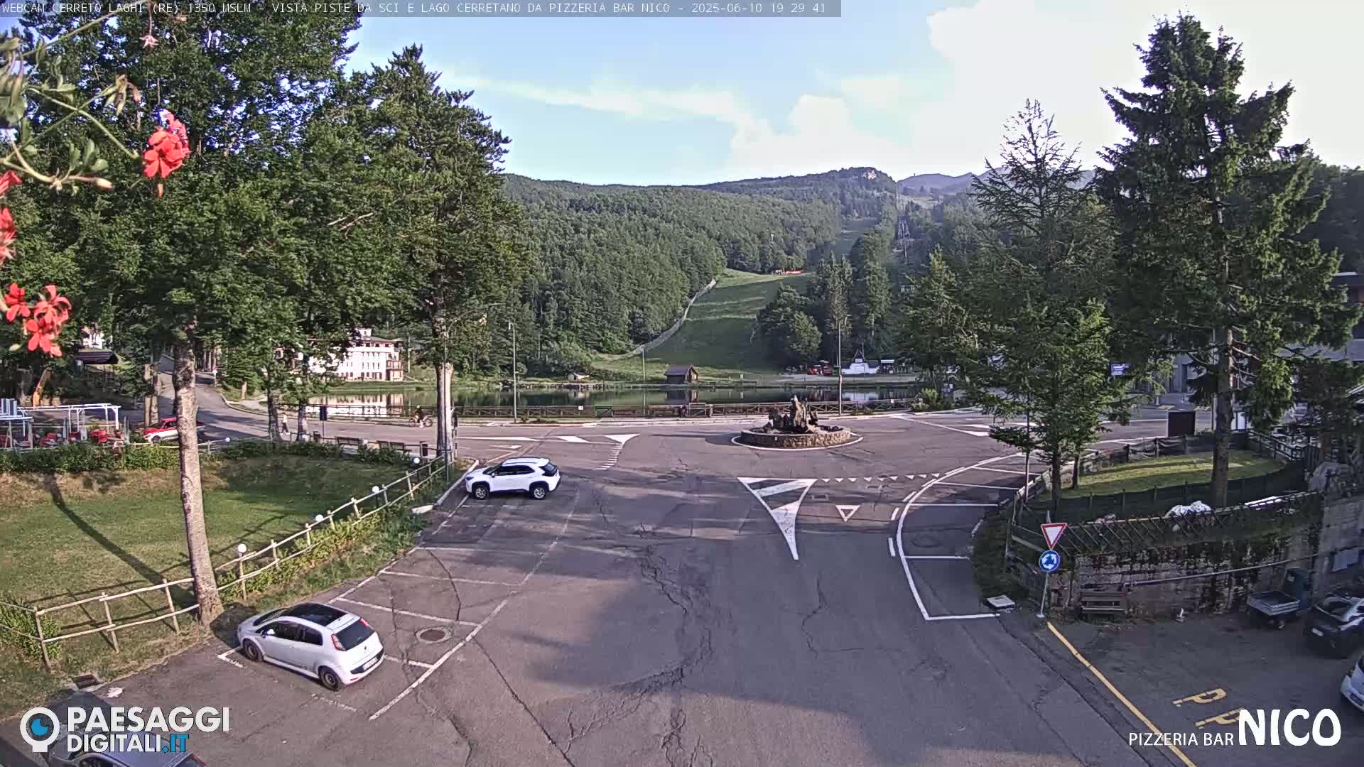 A mostly sunny outdoor scene shows a parking lot, a roundabout with a small fountain, a lake, and a hillside covered in green trees and a ski lift.