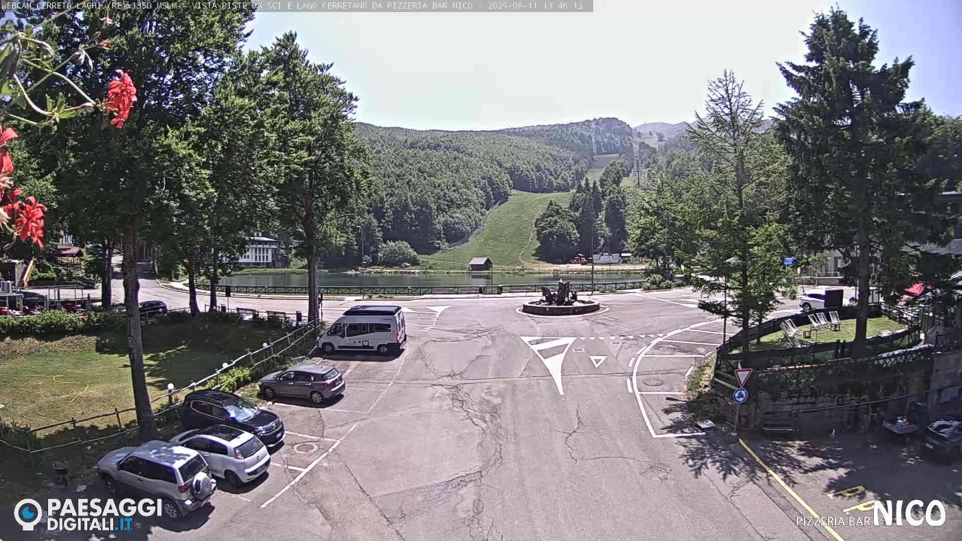 A sunny parking lot overlooks a small lake and a grassy hillside with ski slopes in the background.