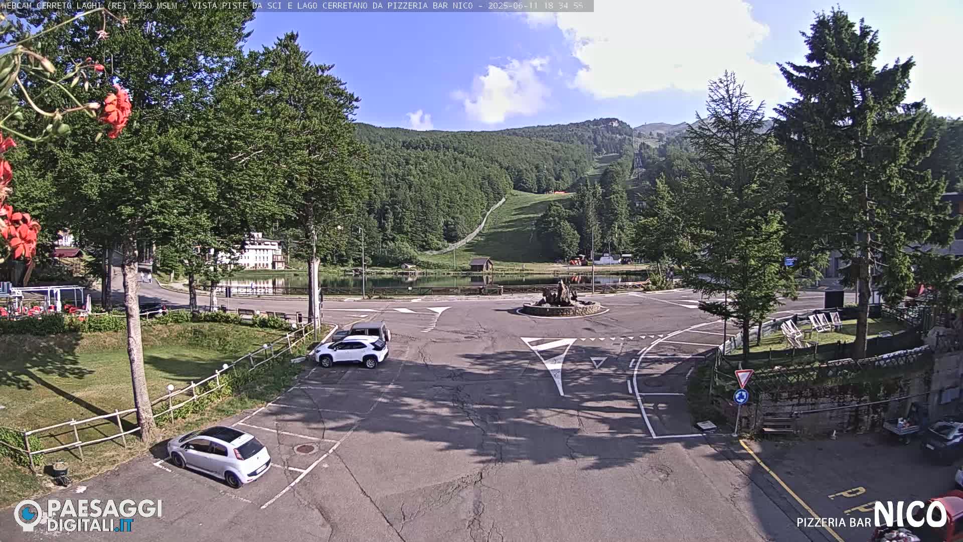A mostly sunny parking lot overlooks a small lake and a ski slope in a mountainous region.