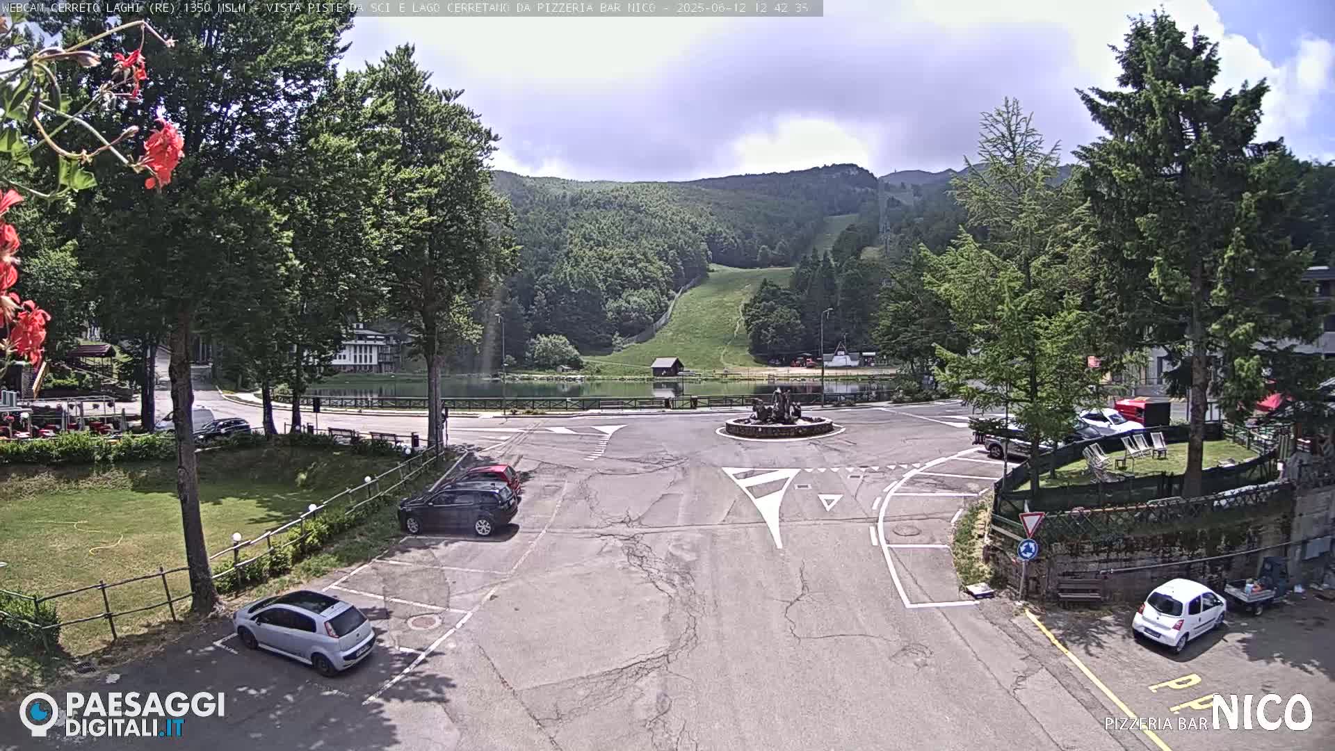A parking lot with several cars is situated in front of a small lake and green mountains under a partly cloudy sky.