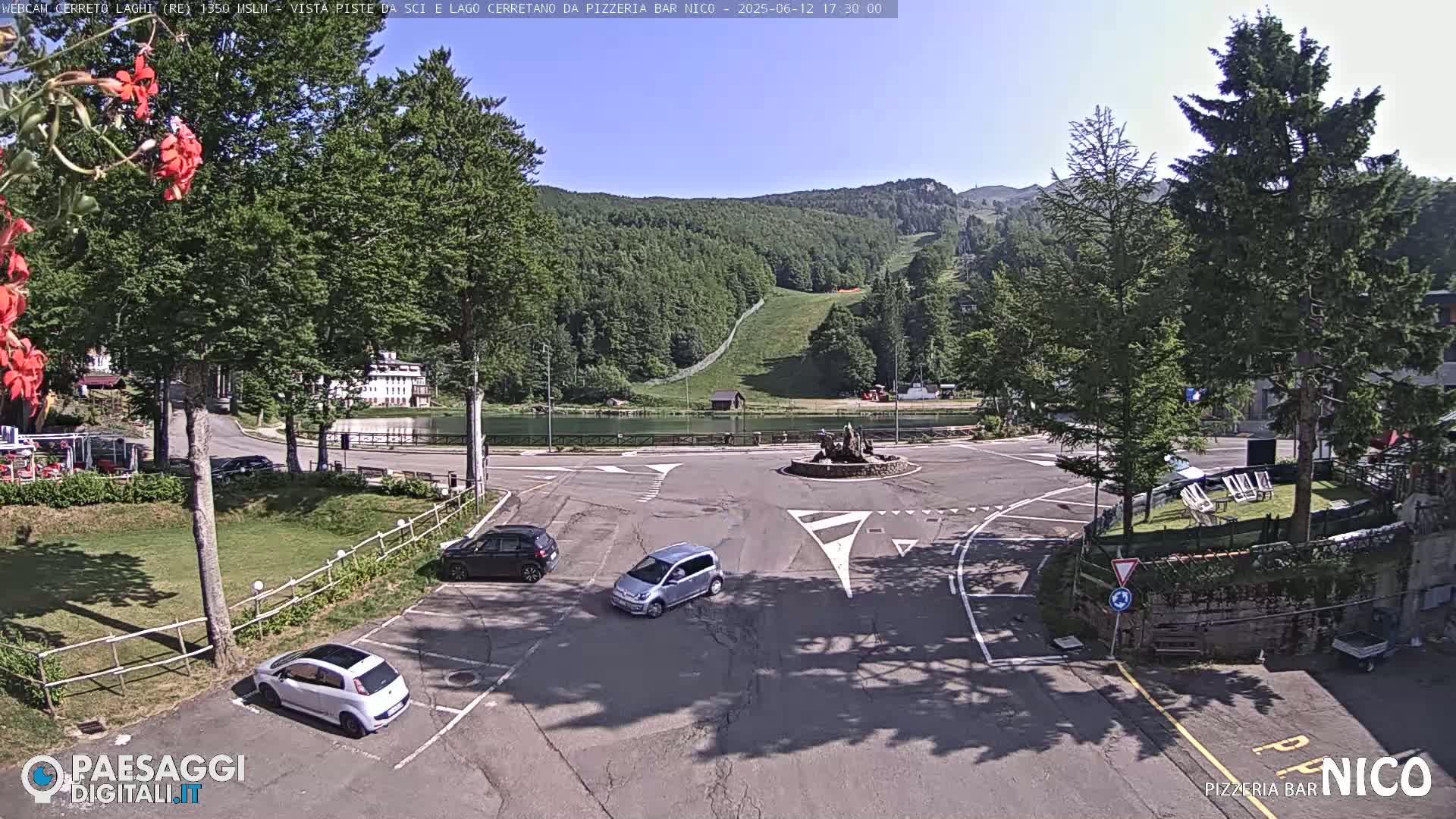 A sunny day overlooking a parking lot, roundabout, small lake, and ski slope in a mountainous region.