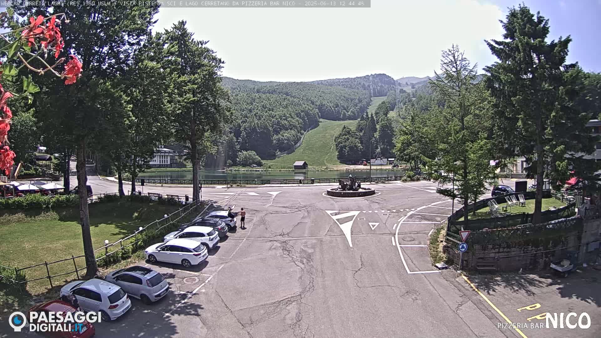 A sunny day at a mountain resort with a small lake, parking lot, and several cars, viewed from above.