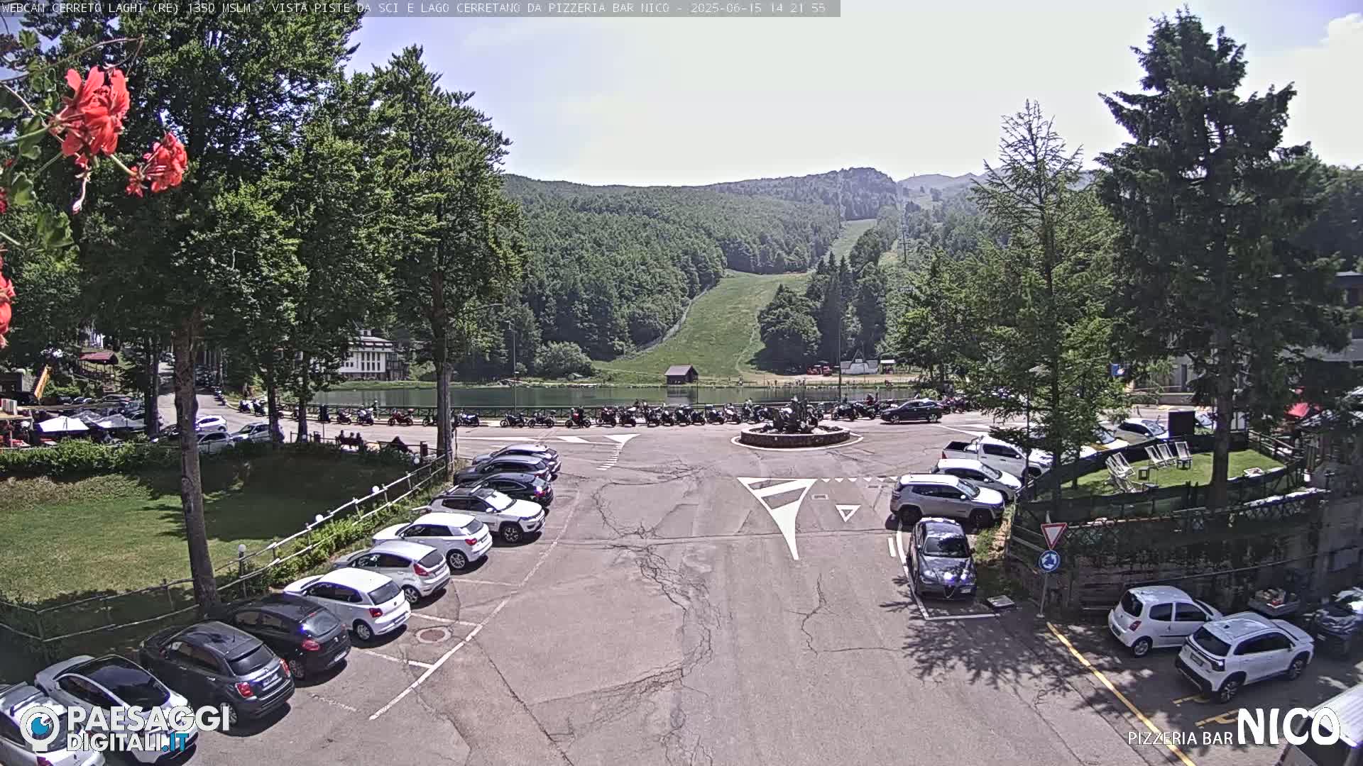 A parking lot with many cars is situated near a calm lake and a mountain with a ski slope, under a mostly sunny sky.