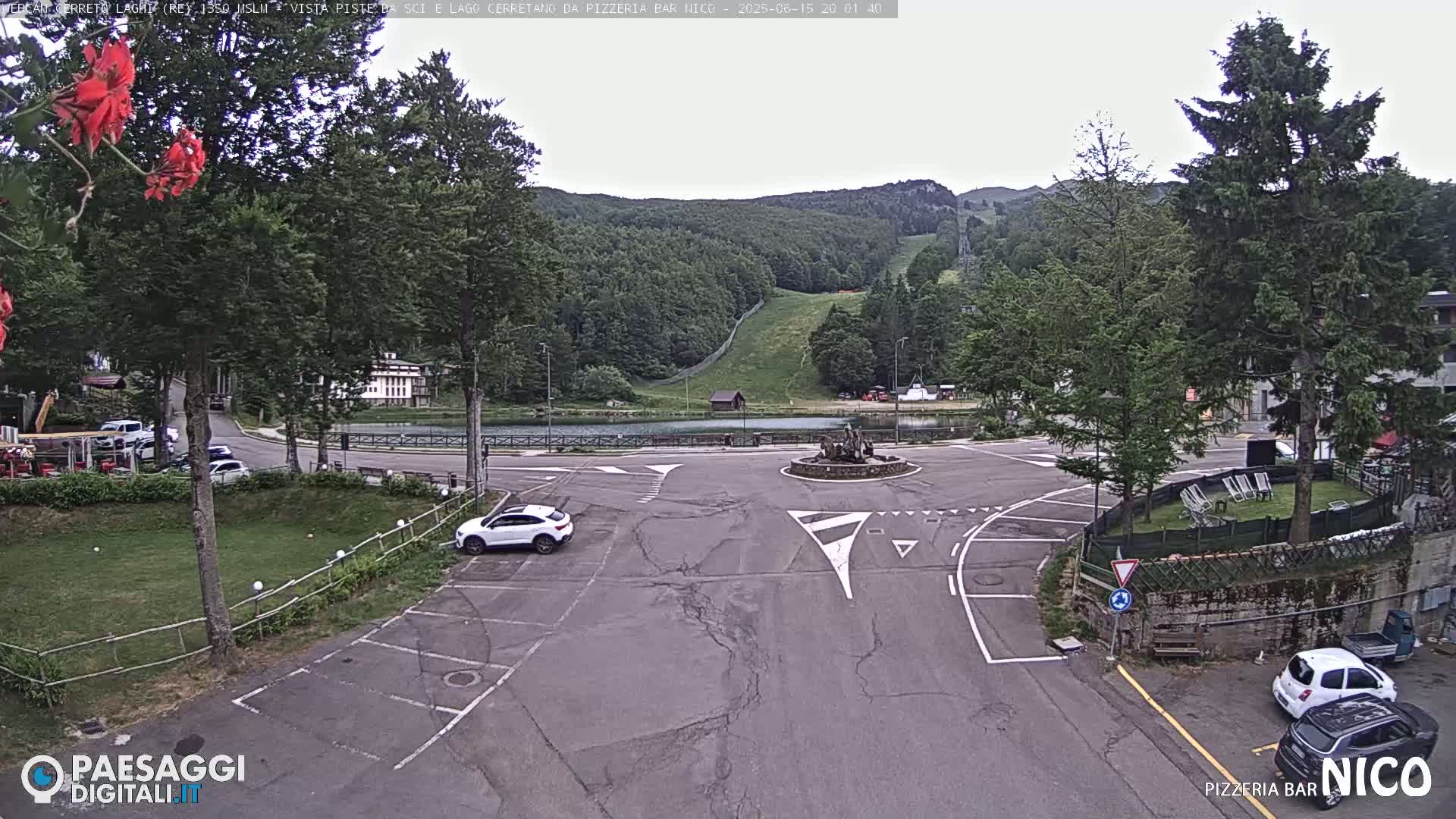 A mostly empty parking lot and roundabout are situated in front of a small lake and a forested hillside under an overcast sky.