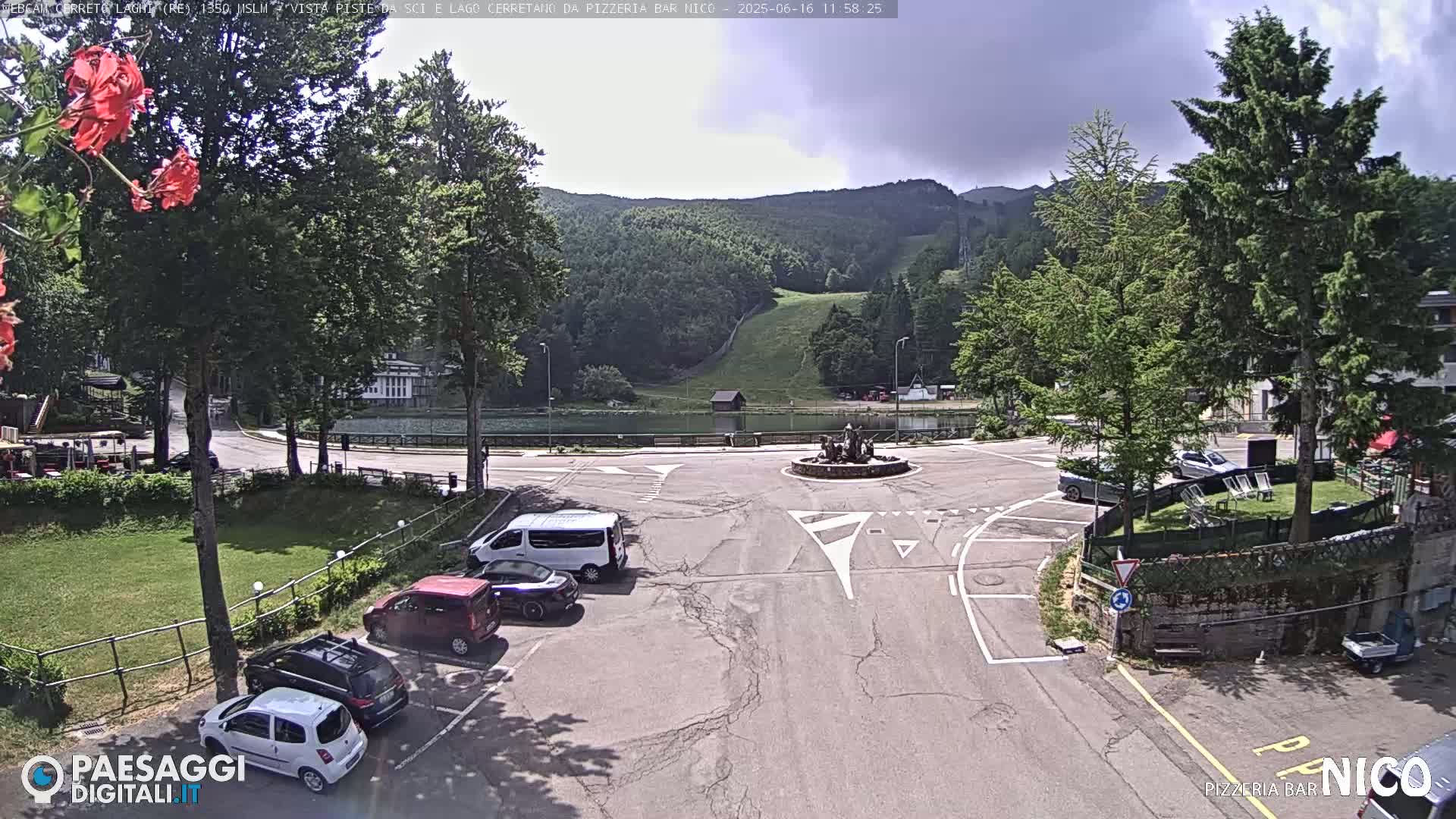 A parking lot with several cars is situated near a small lake and a grassy hill under a partly cloudy sky.
