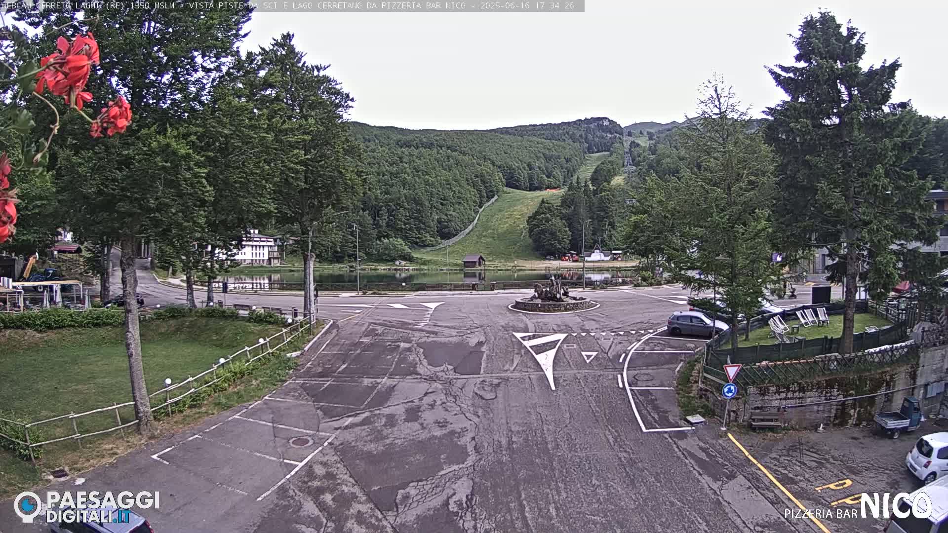 A parking lot and roundabout overlook a small lake, ski slope, and forested mountain under an overcast sky.