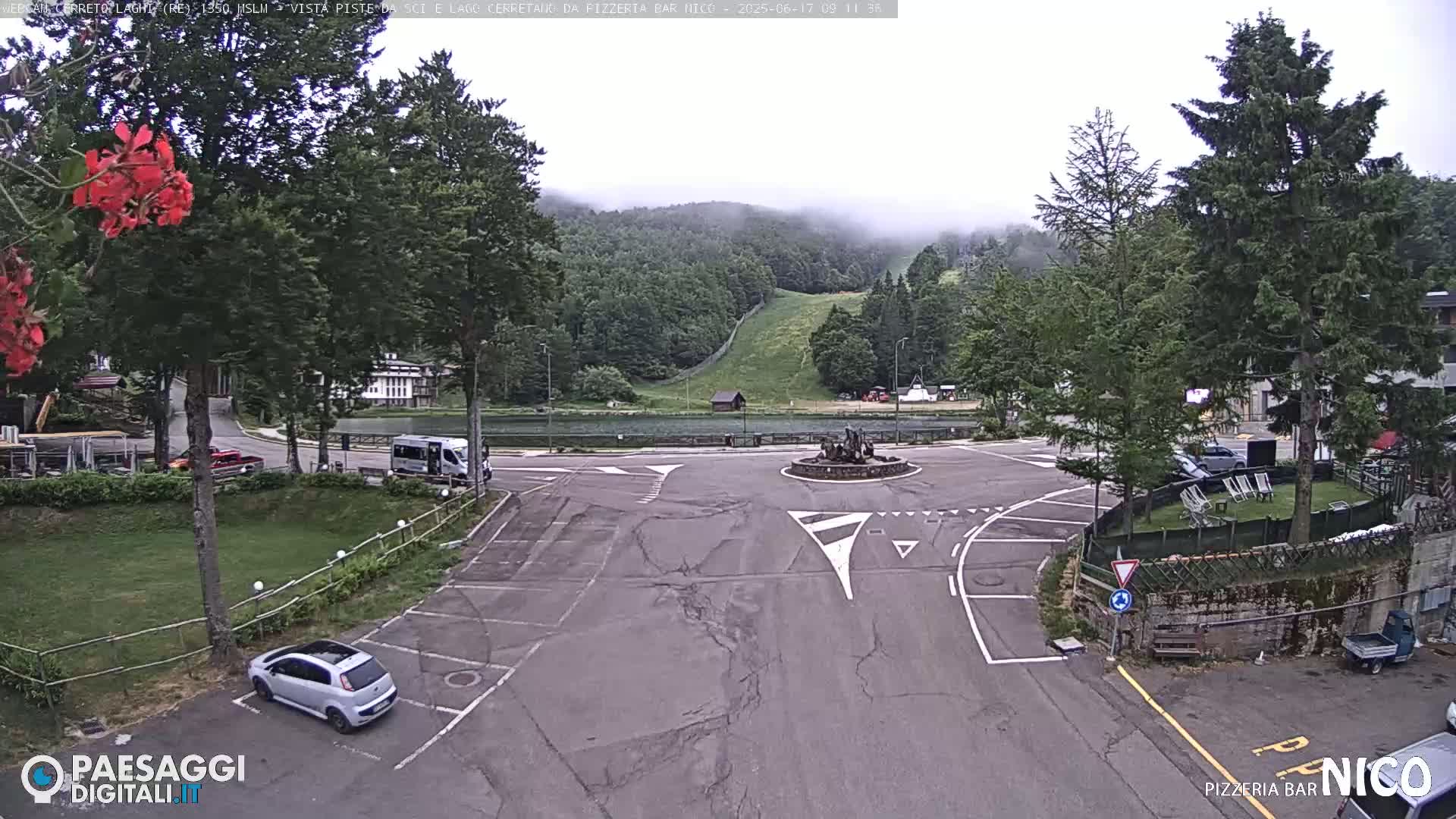 A mostly cloudy day shows a parking lot and roadway intersection near a lake and forested hillside.