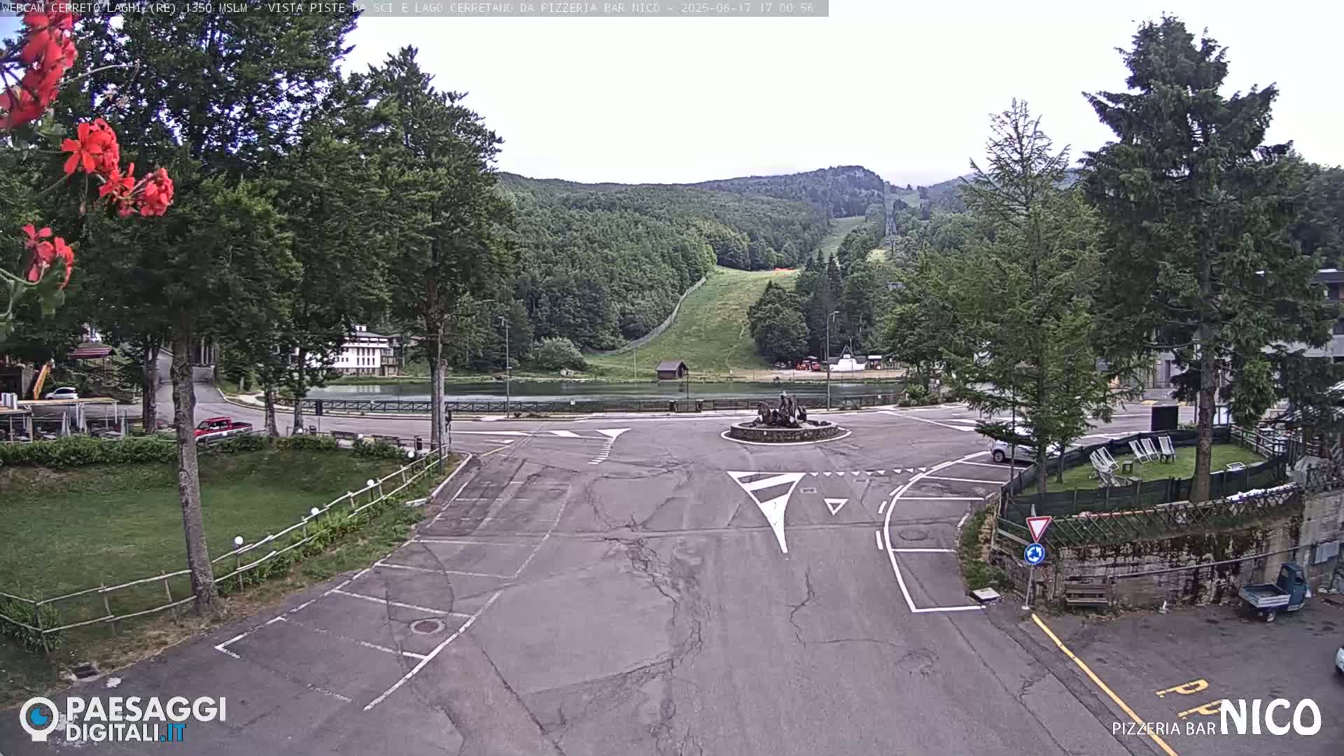 A mostly empty parking lot and roundabout are situated in front of a small lake and a hillside covered in green trees, under an overcast sky.