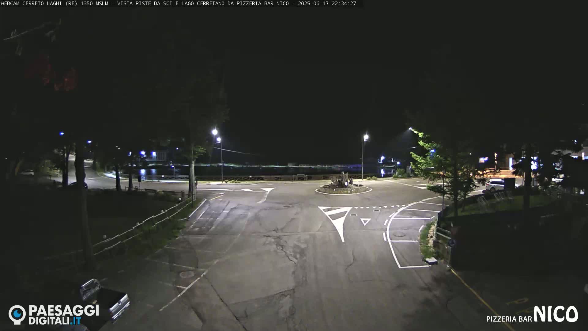 A nighttime view of a mostly empty parking lot and intersection near a body of water, illuminated by streetlights.