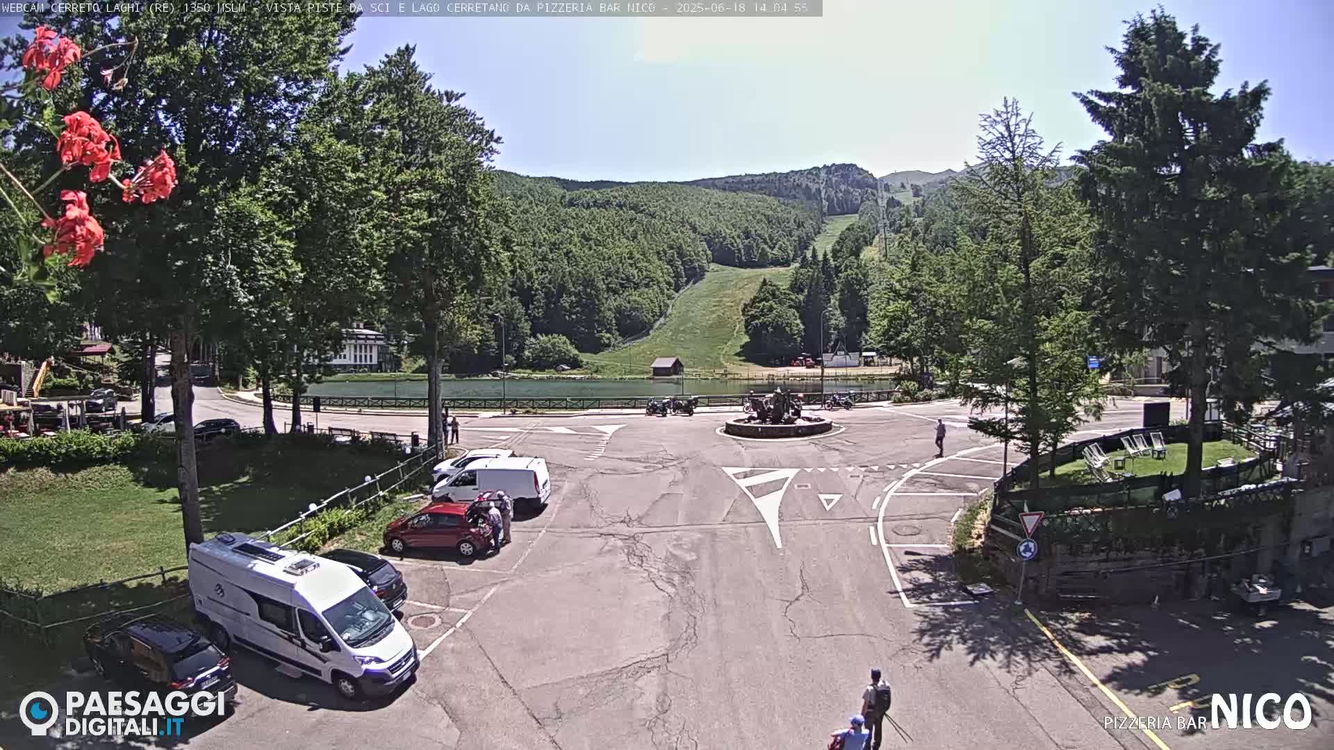 A parking lot with several cars and vans is situated near a small lake and green hills under a sunny sky.
