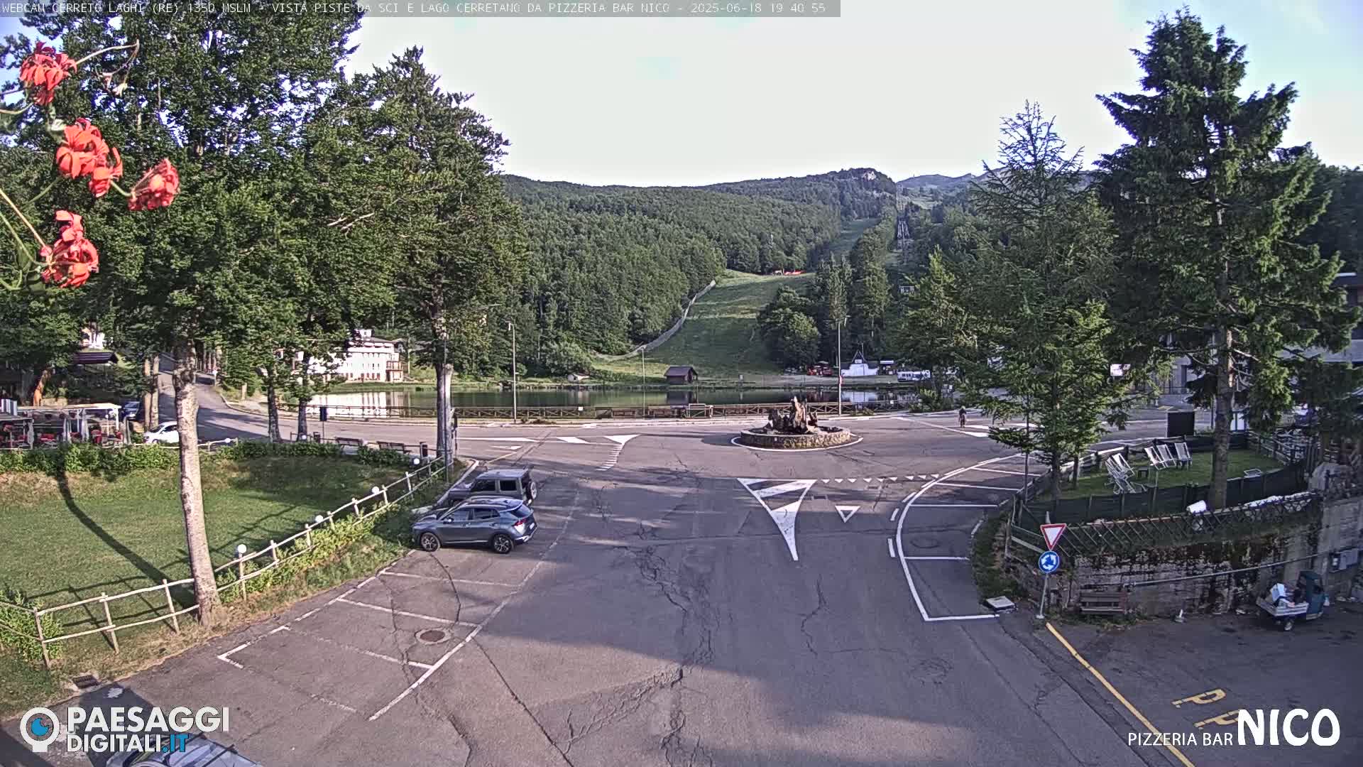 A mostly sunny view of a parking lot and roundabout near a lake and ski slope, with several cars parked and surrounded by trees.