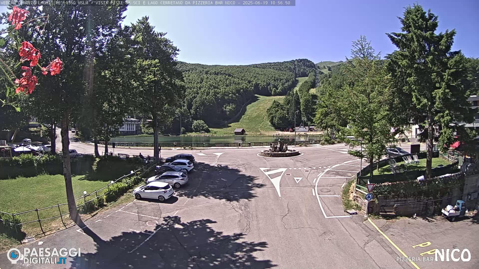 A sunny, outdoor view of a parking lot with several cars, a small lake, and a grassy hill in the background.