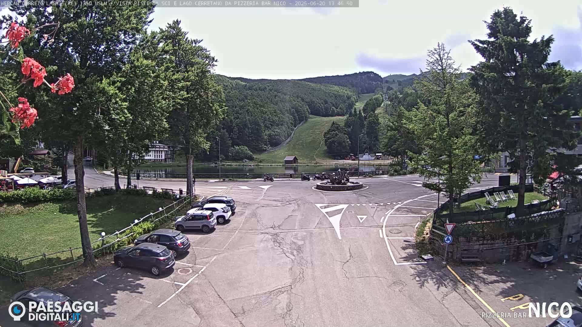 A parking lot with several cars is situated near a roundabout and a small lake, with a hill covered in lush green trees and a ski slope in the background under a partly cloudy sky.