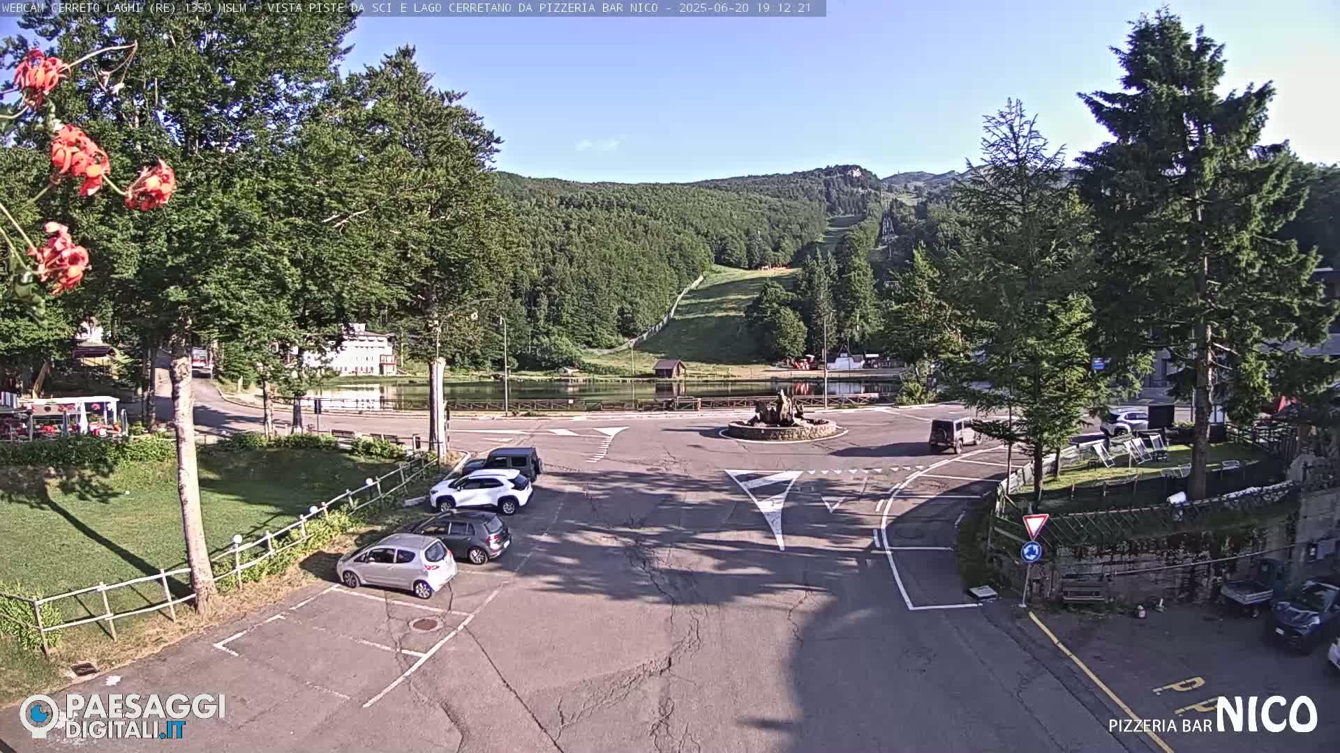 A sunny day reveals a parking lot with several cars near a roundabout and a small lake, backed by a forested hillside with a ski slope visible in the distance.