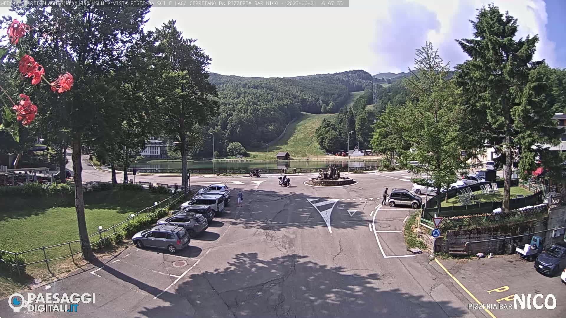 A parking lot with several cars is situated near a small lake and a roundabout, with a background of green hills and a ski slope under a partly sunny sky.
