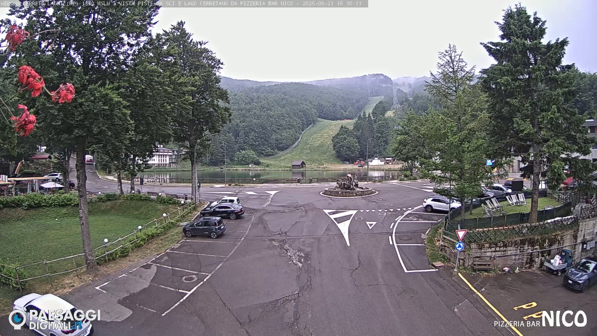 A parking lot with several cars is situated near a small lake and a ski slope in the background, under an overcast sky.