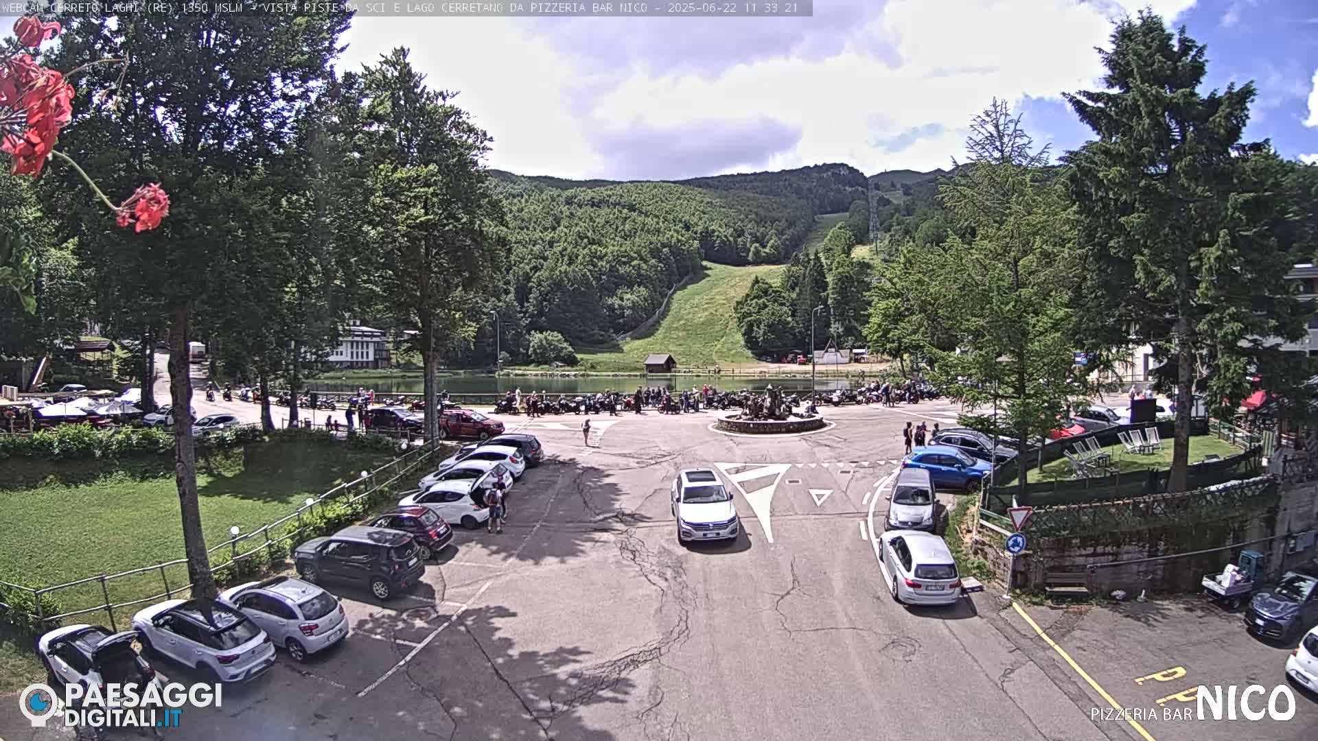 A parking lot with cars is next to a lake and a small crowd of people, under a partly cloudy sky, with hills and trees in the background.