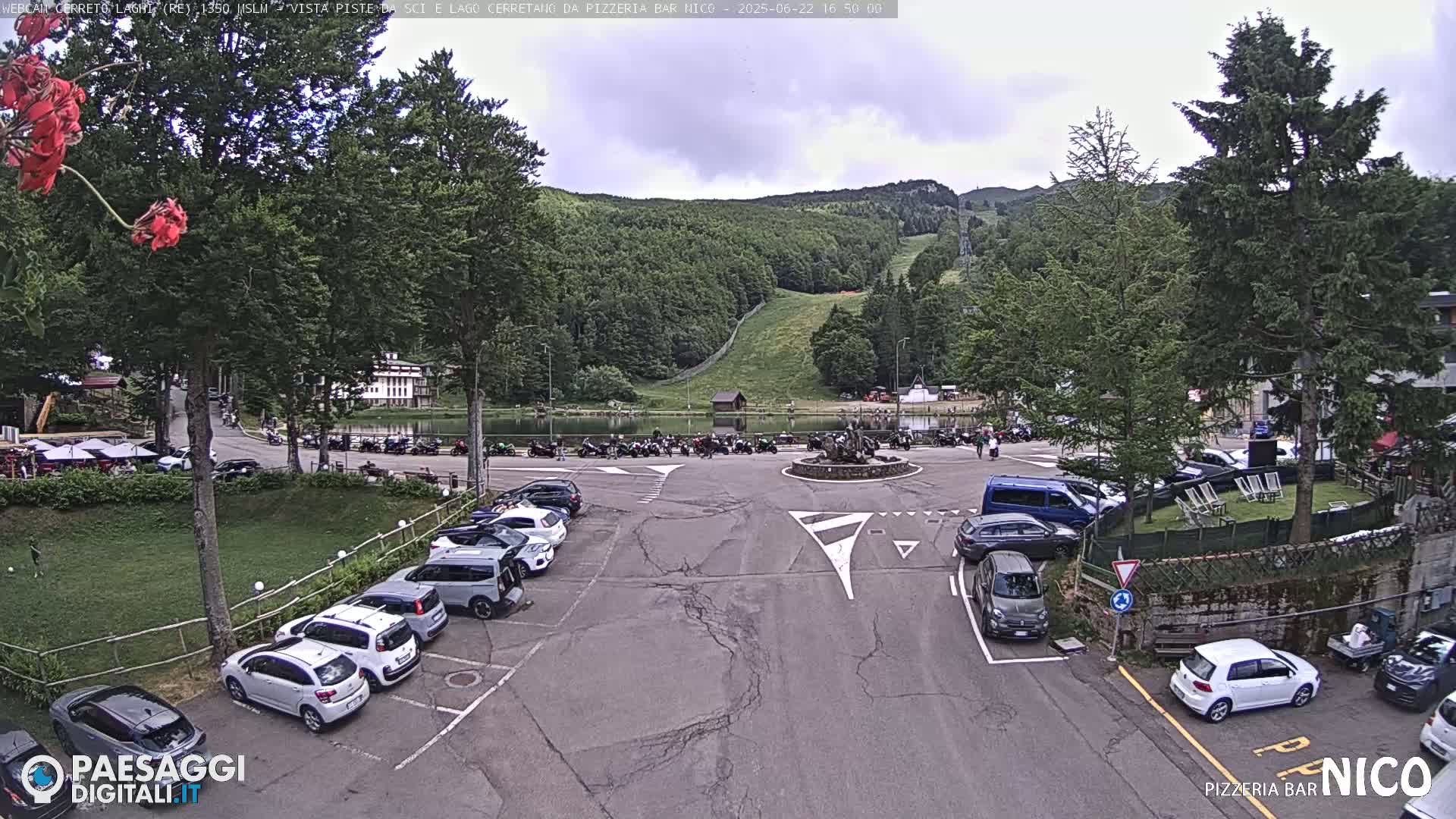 A parking lot with cars surrounds a small lake and a mountainside with a ski lift, under a partly cloudy sky.