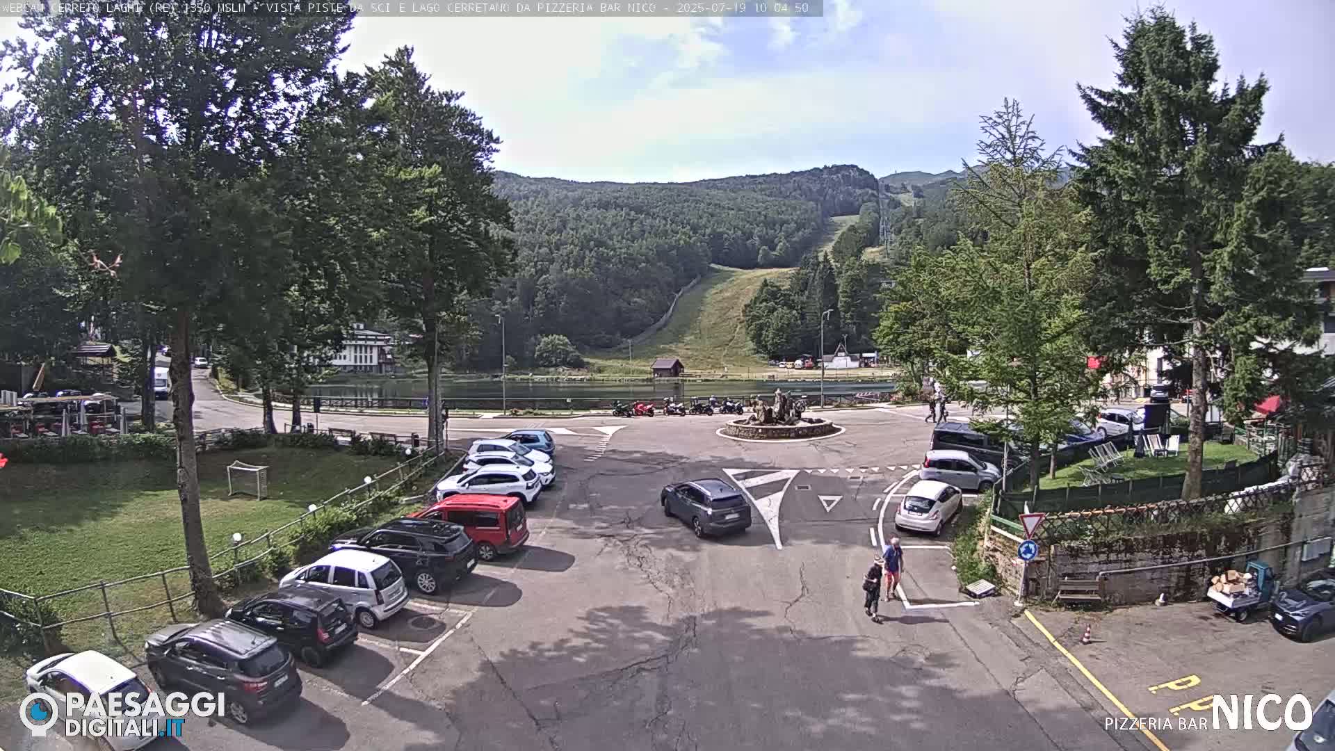 A sunny day shows a parking lot filled with cars surrounding a small lake and roundabout, with a hill covered in trees and a ski slope in the background.